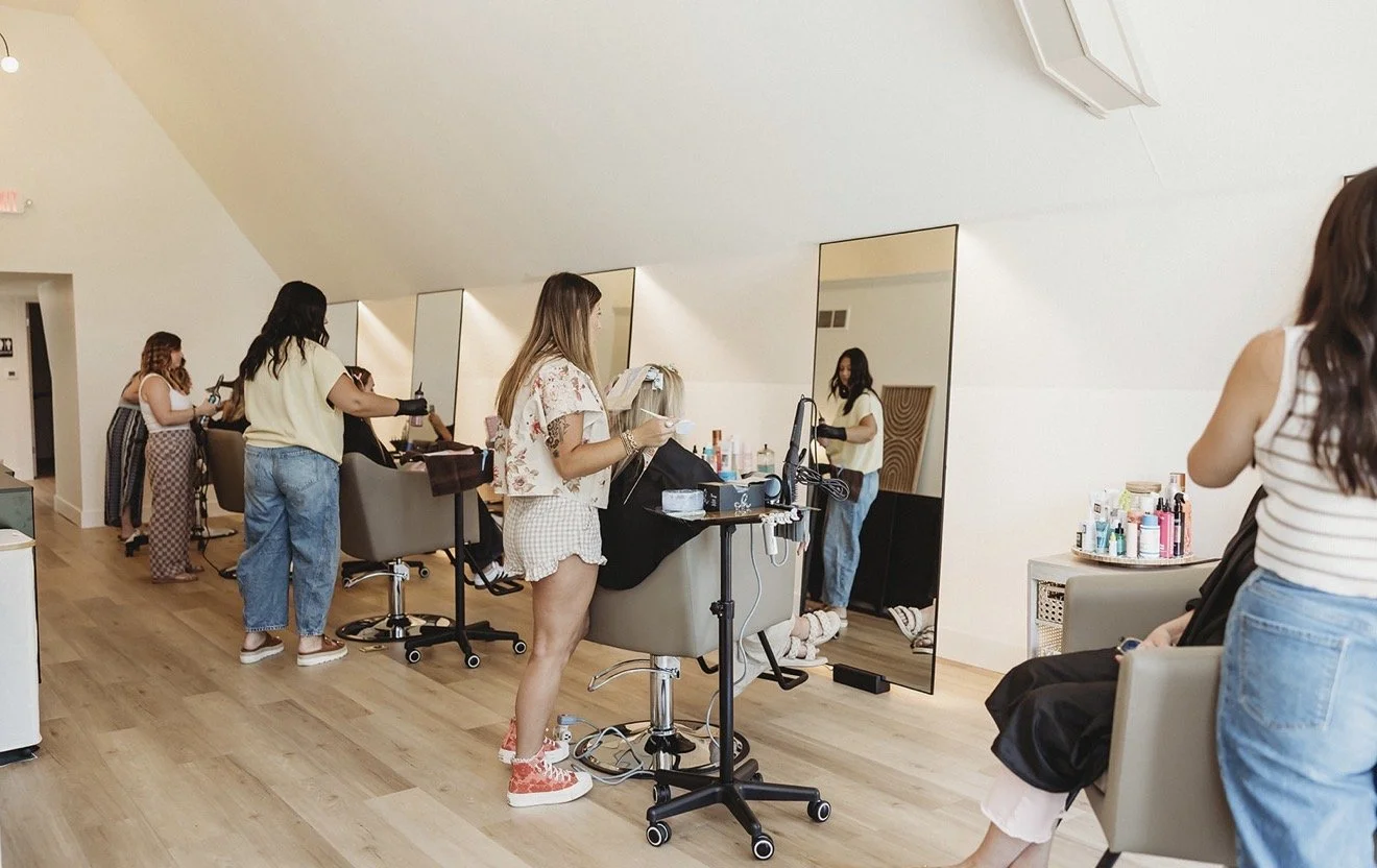 Hair stylists working with clients in a bright salon lined with mirrors and styling stations