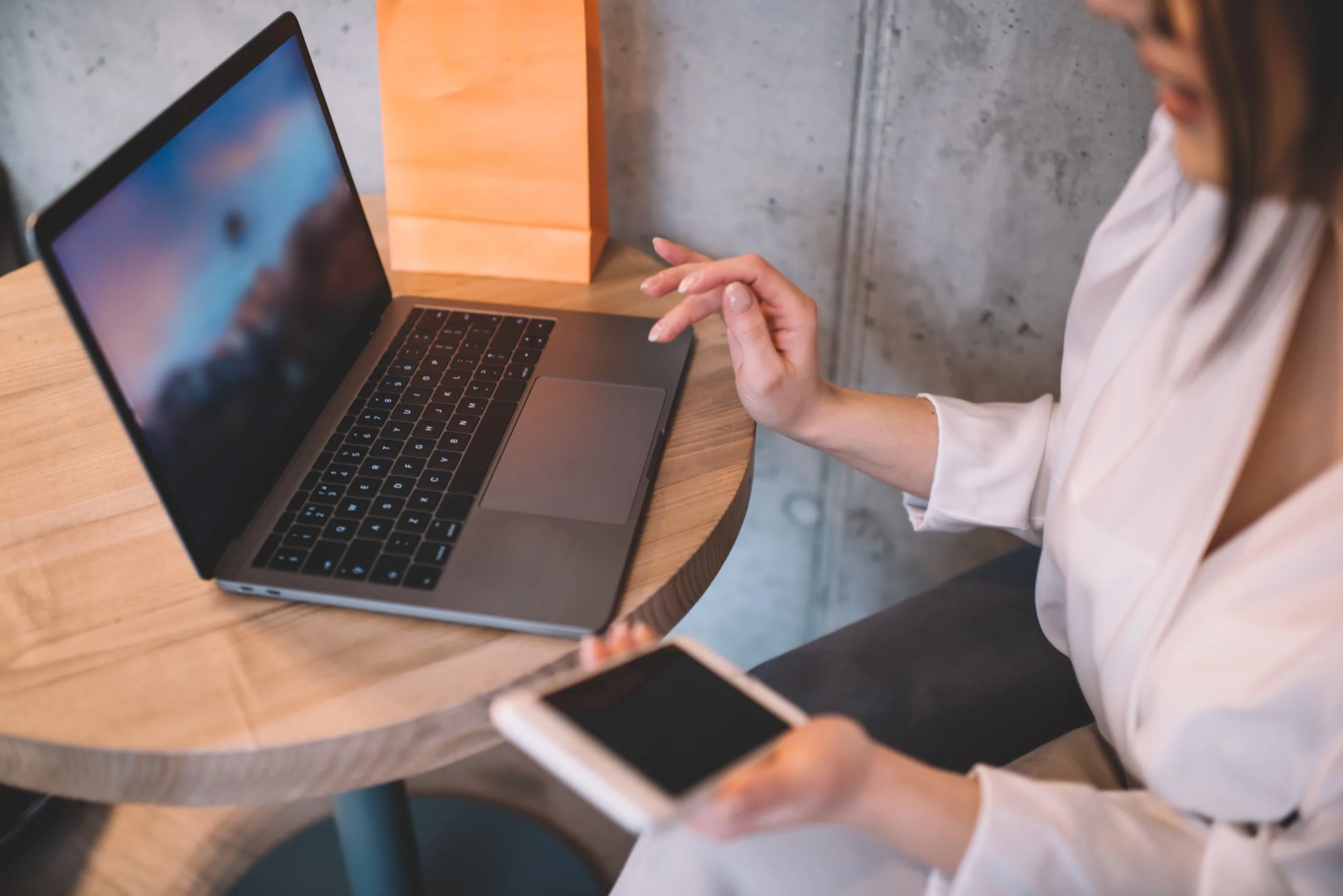 A person sitting at a wooden table using a laptop and holding a smartphone.