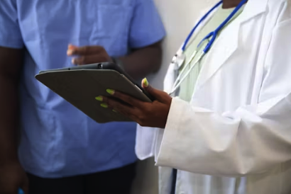 A healthcare professional in a white coat with a stethoscope around their neck holding a tablet, with another person in scrubs in the background.
