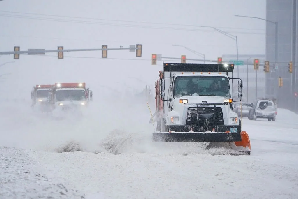 Snowplow clearing snow on a city street during a snowstorm, with traffic lights and vehicles in the background.