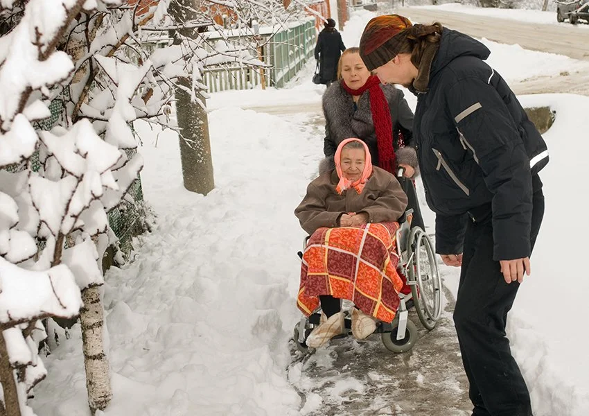 An elderly woman in a wheelchair wrapped in a colorful blanket, with a young woman and a man standing nearby, outdoors on a snowy day.