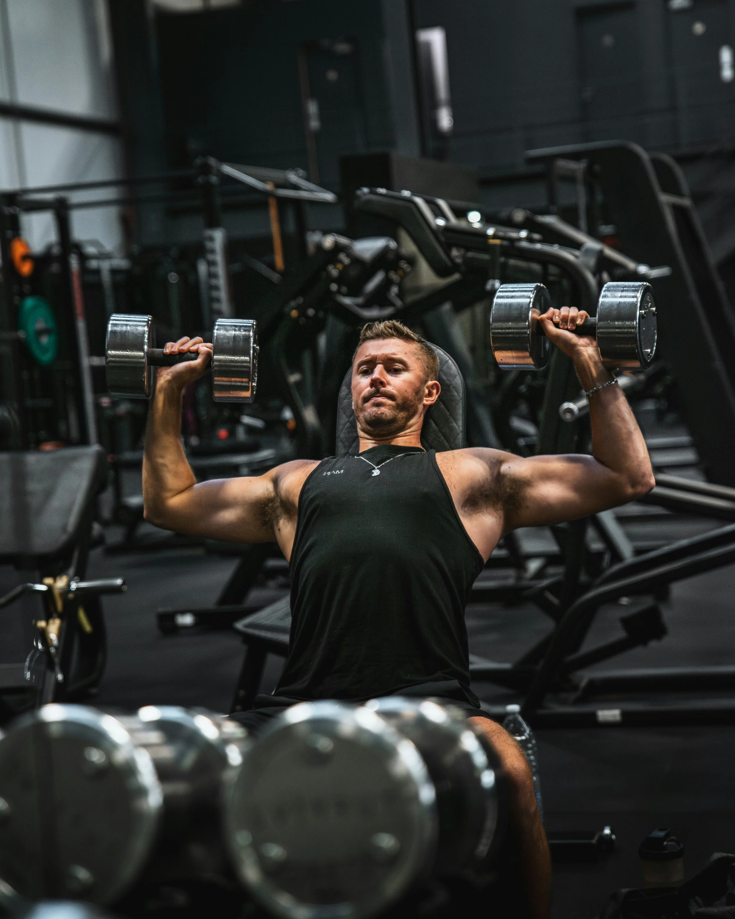 A man exercises in a gym, lying on a weight bench and lifting dumbbells. The gym has various workout equipment including barbells and weight plates in the background.