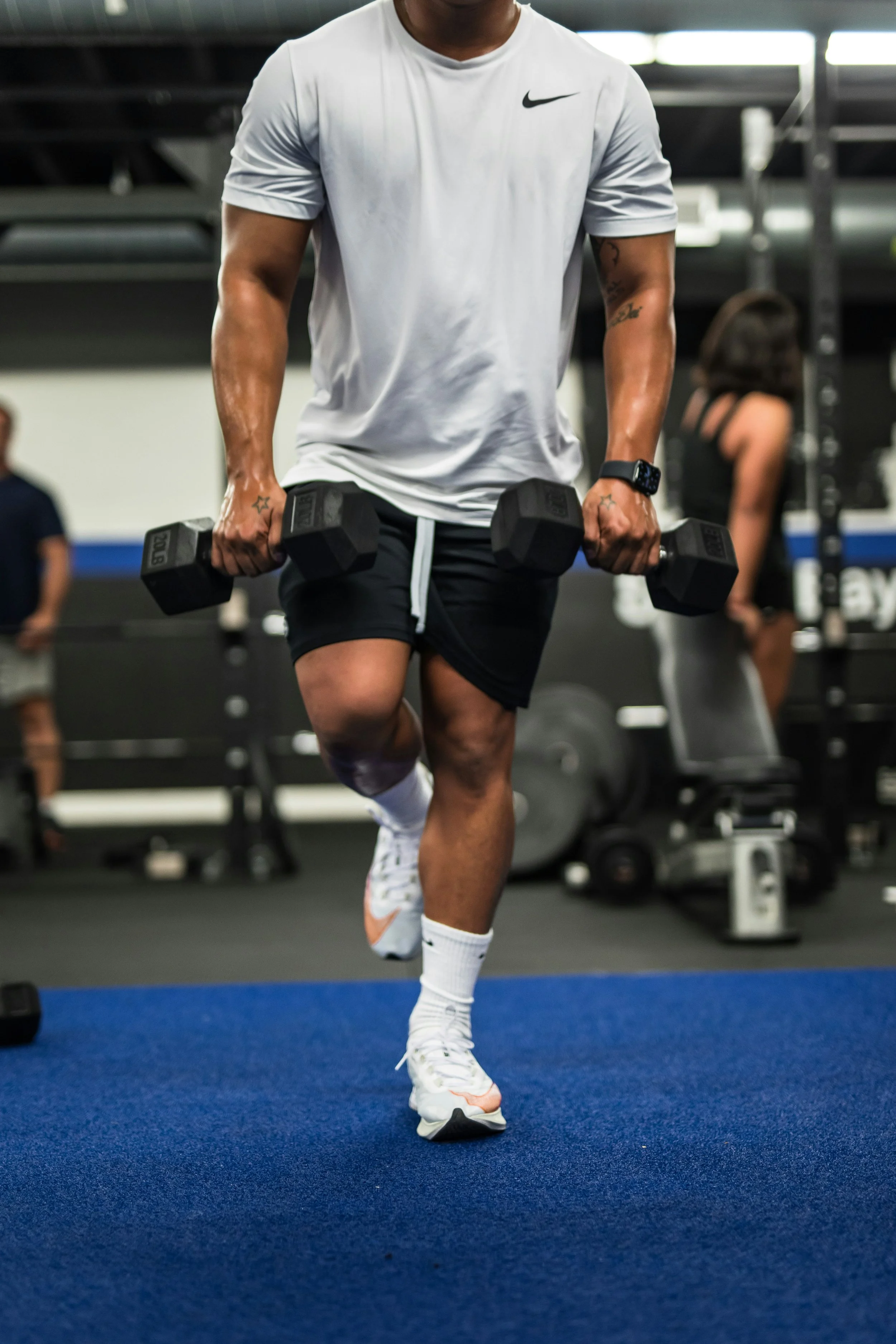 A person in athletic clothing doing a workout in a gym, lifting dumbbells with both hands, with gym equipment and other people in the background.