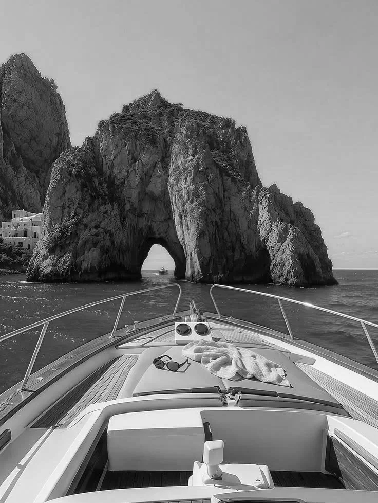 View from the bow of a boat looking towards a large rock formation with a natural arch in the water, with towels and sunglasses on the boat's deck.