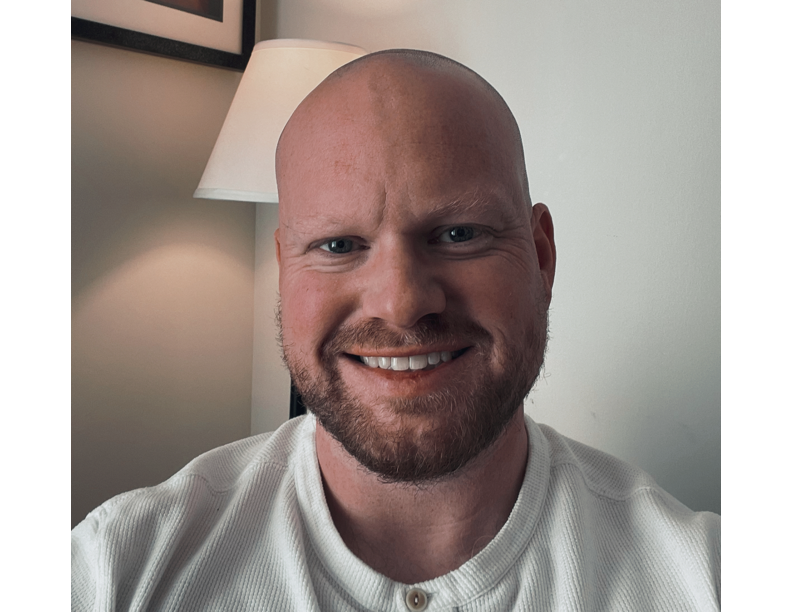 A smiling man with a beard and head shaved, wearing a white shirt, sitting in a room with a lamp and picture on the wall in the background.
