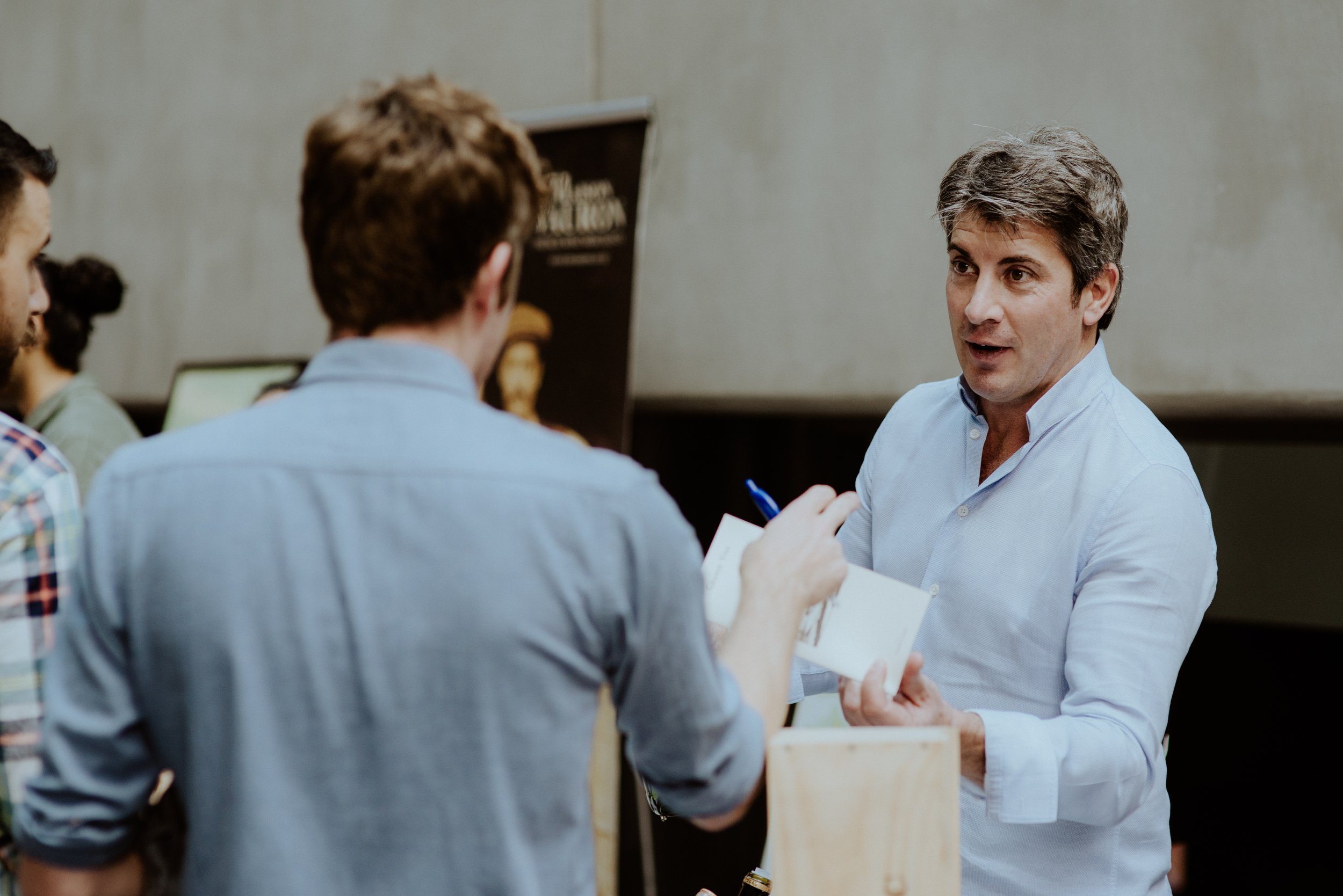 A man with gray hair and a blue shirt engaging in conversation with two younger men with brown hair, one wearing a plaid shirt and the other in a light-colored shirt, in an indoor setting.