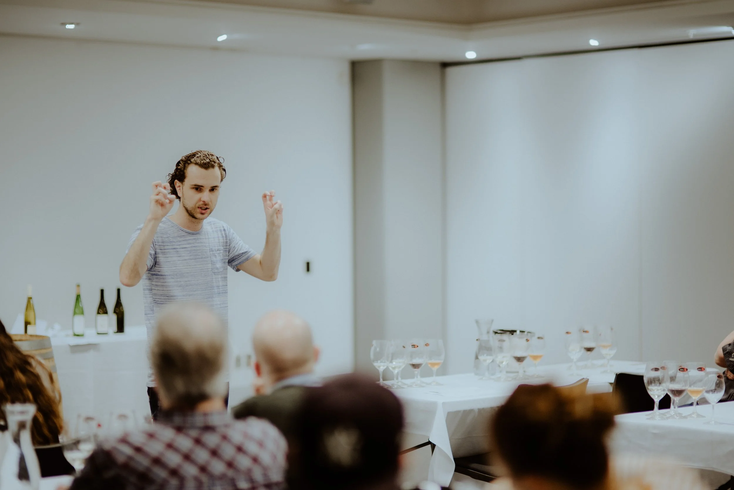 A man (Connal Finlay) with curly hair sitting on a chair in a room with tables and bottles, appearing to speak or perform in front of an audience during an event with a banner and clock on the wall.