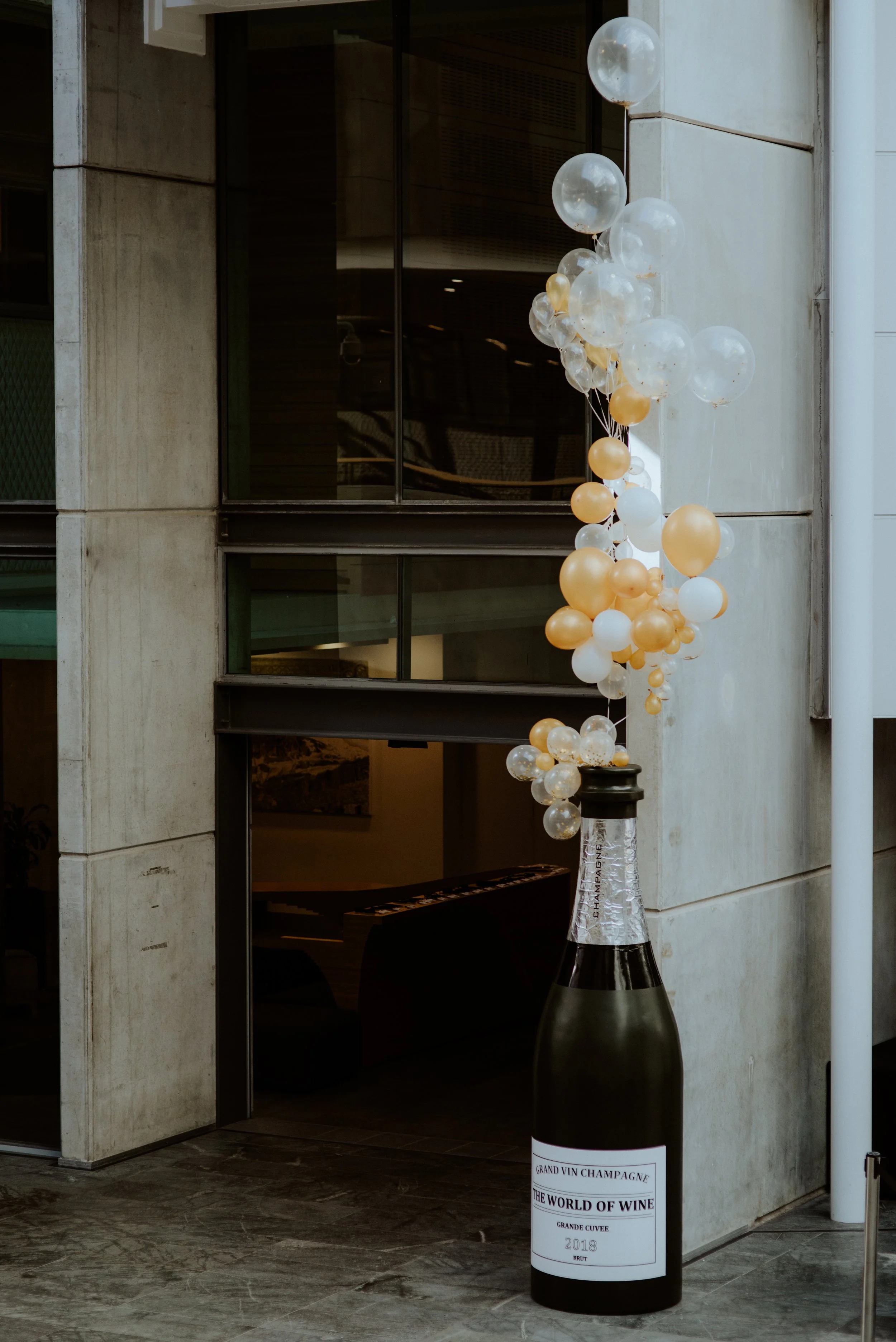 Decorative balloon arrangement placed on a large champagne bottle sculpture outside a building entrance.