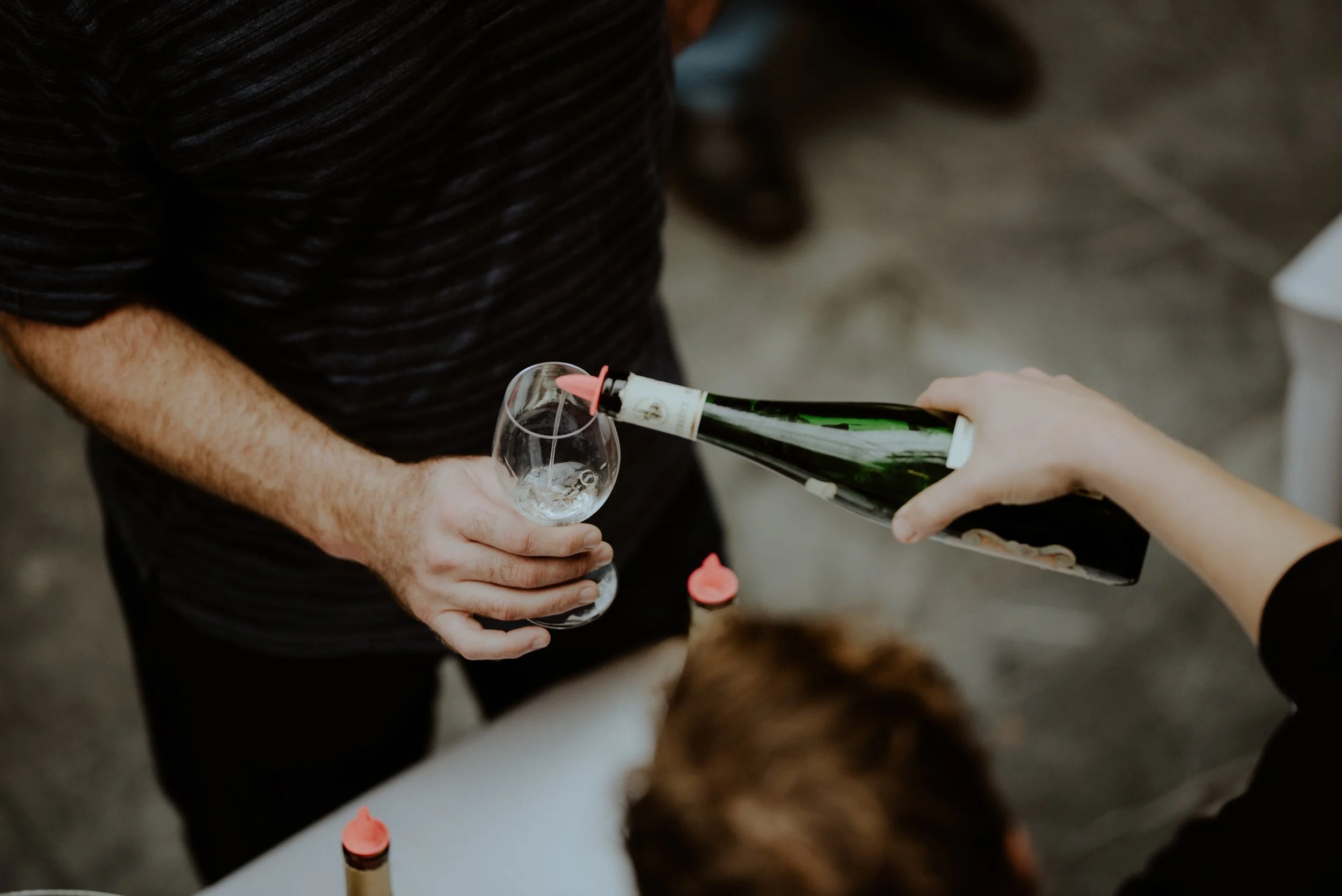 Person pouring white liquid from a wine bottle into a wine glass held by another person.