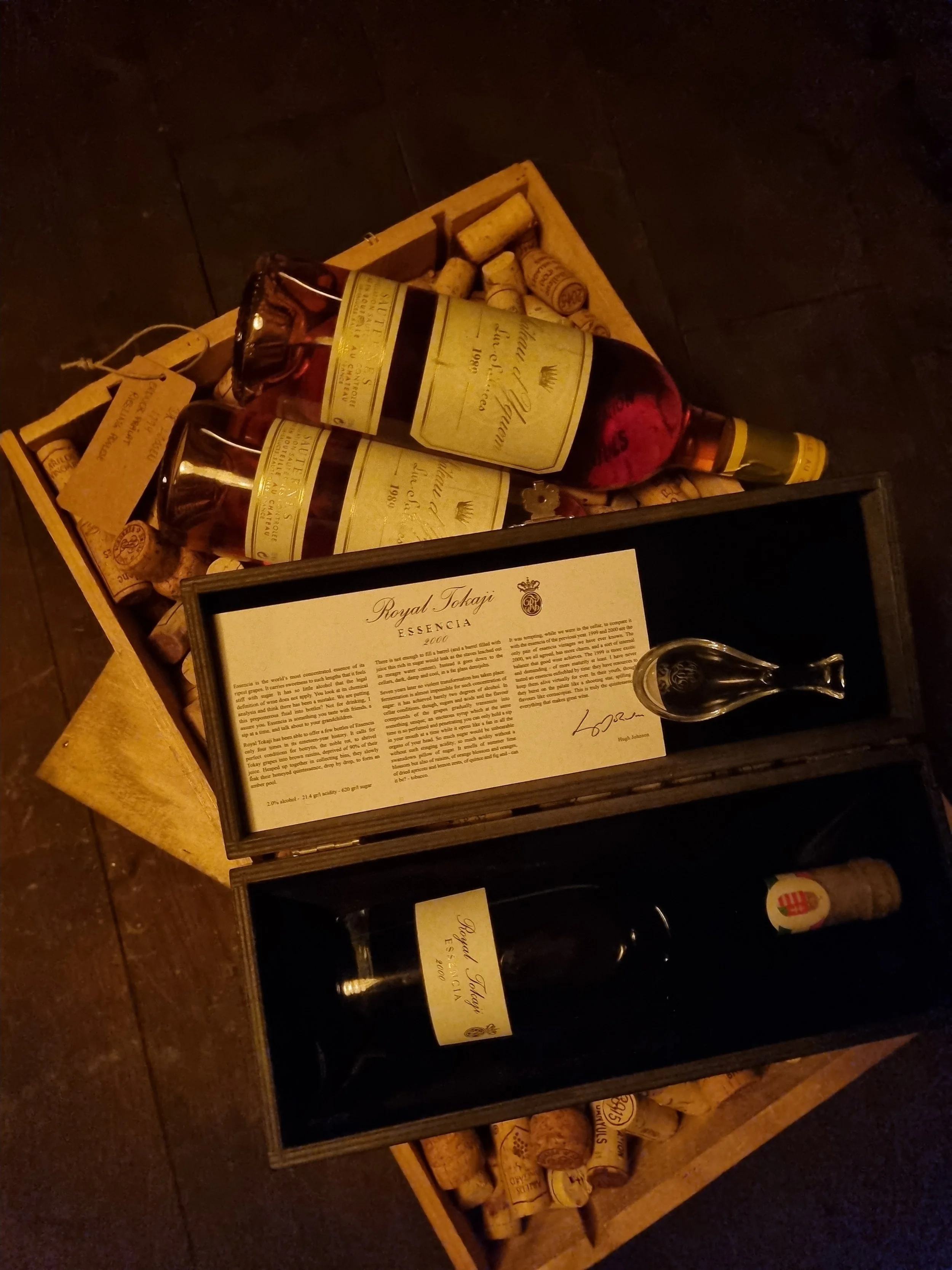 A wooden display shelf with bottles of wine and corks, behind a glass case with a sheet of paper and a wine opener, set on a wooden table.