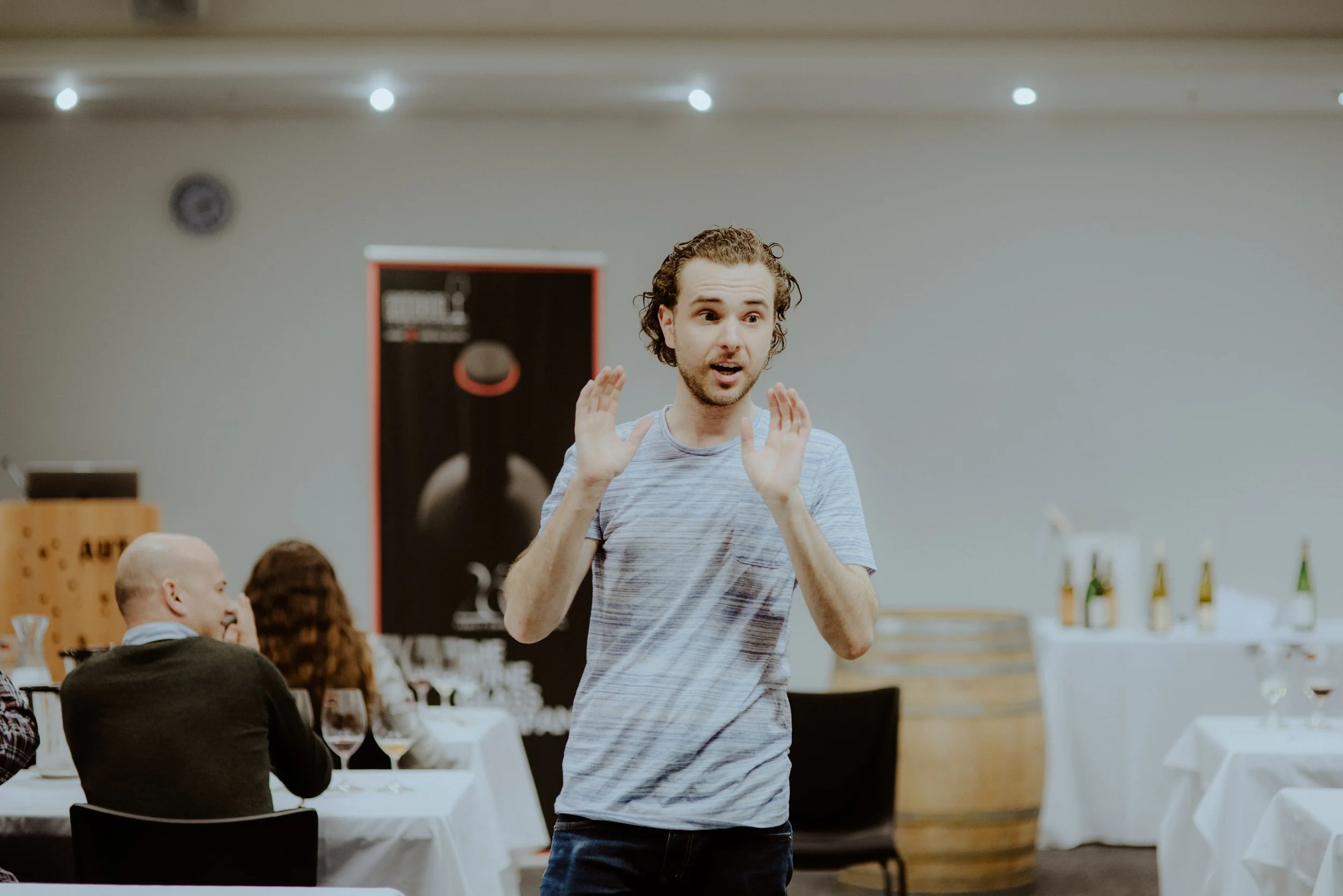 A young man (Connal Finlay) with curly hair and a striped t-shirt appears to be speaking passionately with hand gestures in a social gathering or event, with people seated at tables and bottles on display in the background.