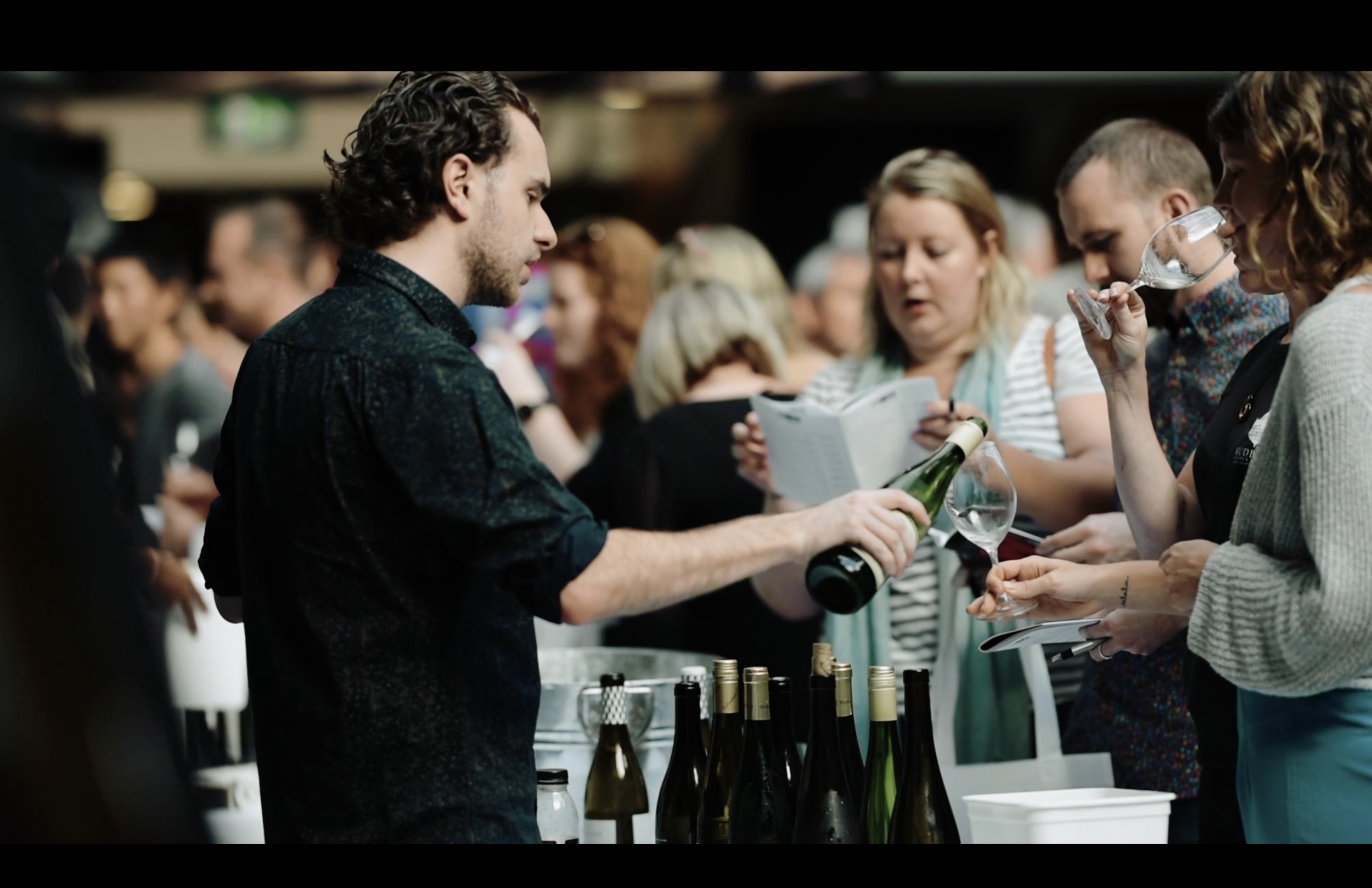 A man (Connal Finlay) pours wine for a woman at a wine tasting event, surrounded by other attendees.