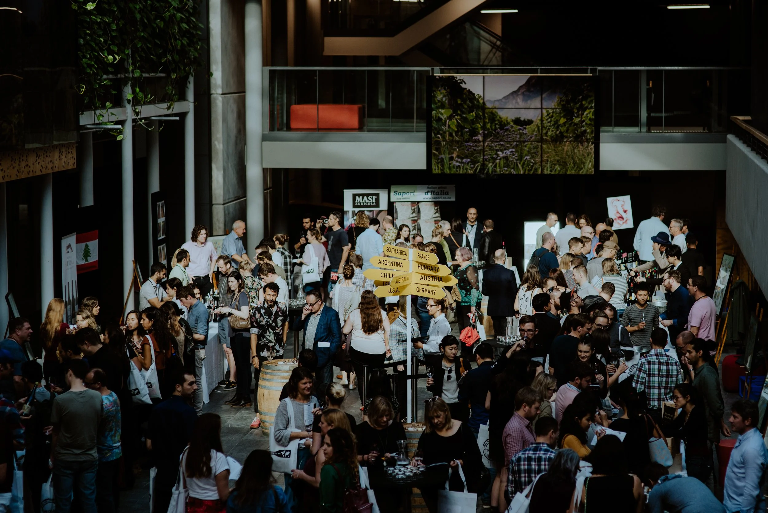 Crowd of people gathered indoors at an event or exhibition, with informational signs and artwork on display.