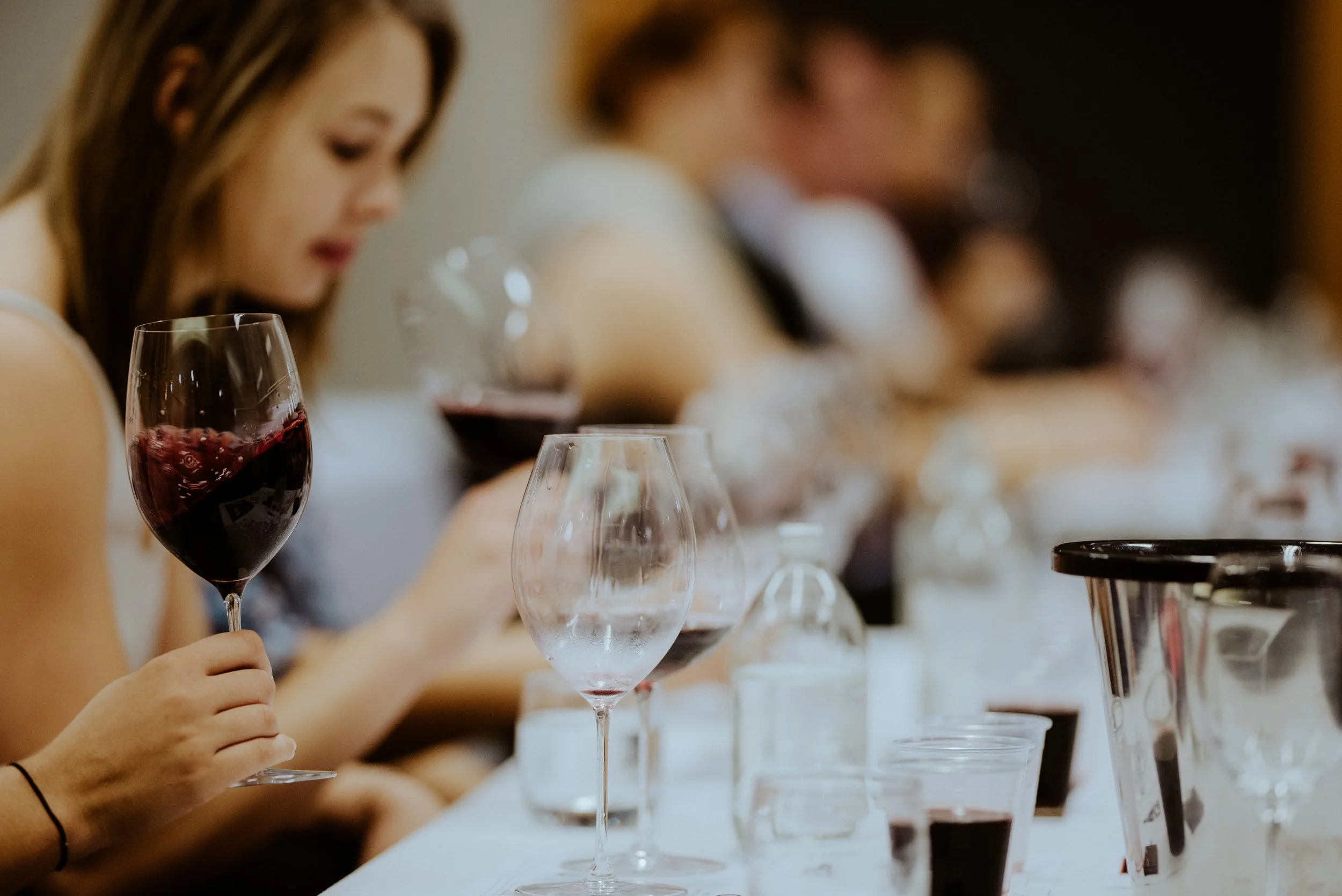 Close-up of a woman with brown hair holding a glass of red wine, with several empty wine glasses and wine bottles on a white table in front of her. The background shows other people toasting with glasses.