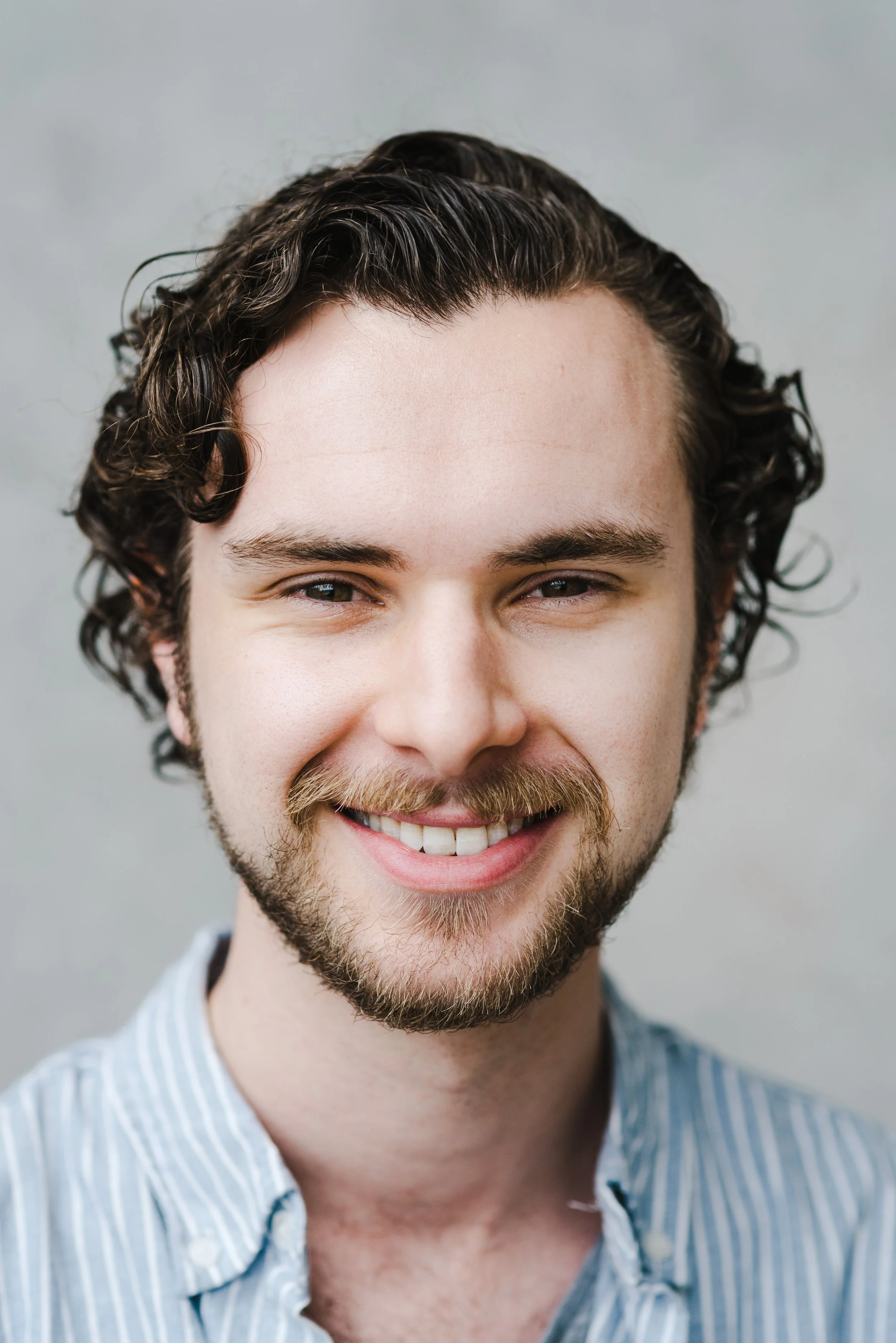 Close-up of a smiling man (Connal Finlay) with curly dark hair, beard, and mustache, wearing a blue and white striped shirt.