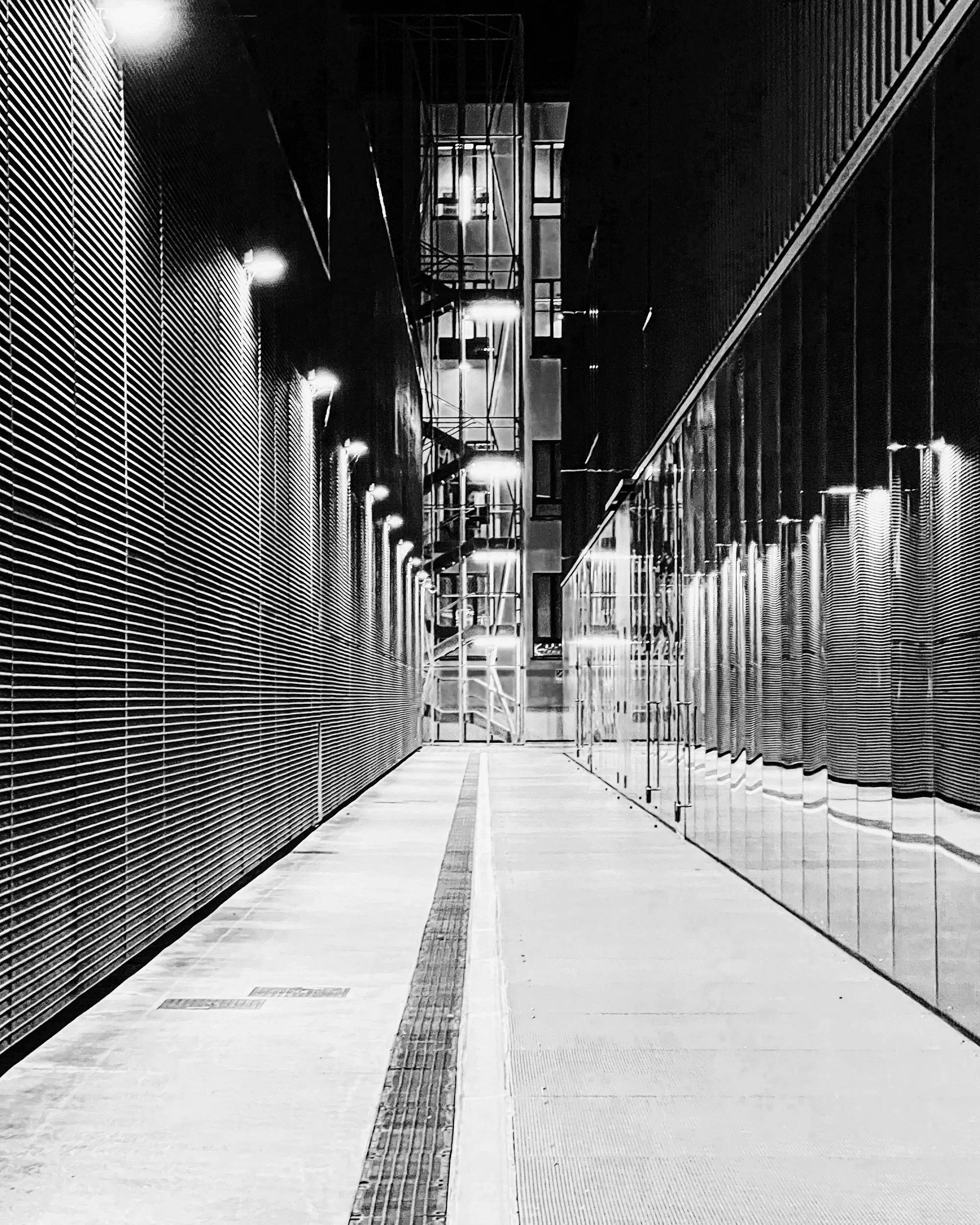 Nighttime view of an empty, well-lit walkway between modern buildings with metallic siding and glass windows.