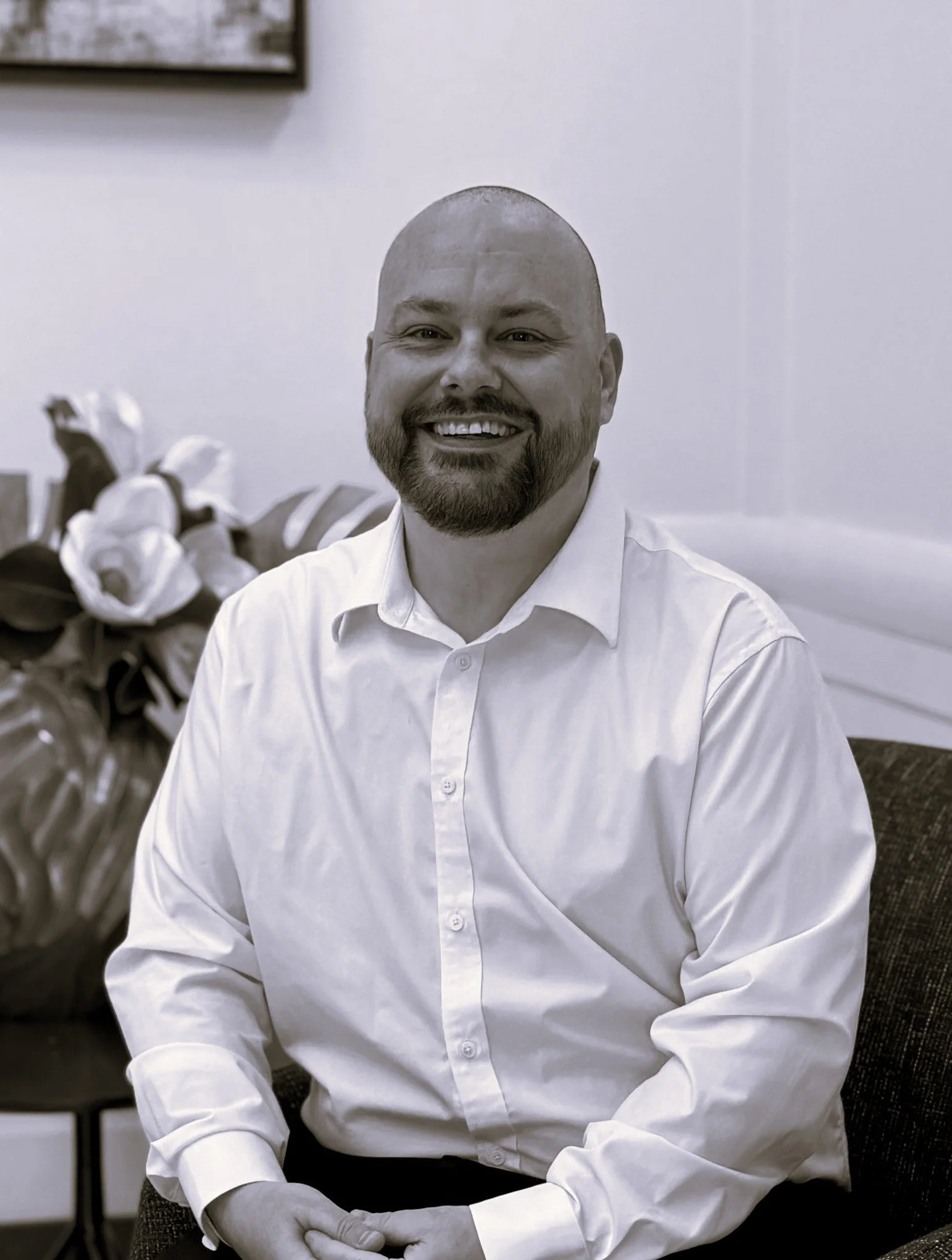 A man with a bald head, beard, and mustache smiling in an office setting, wearing a white dress shirt, with plants and framed artwork in the background.