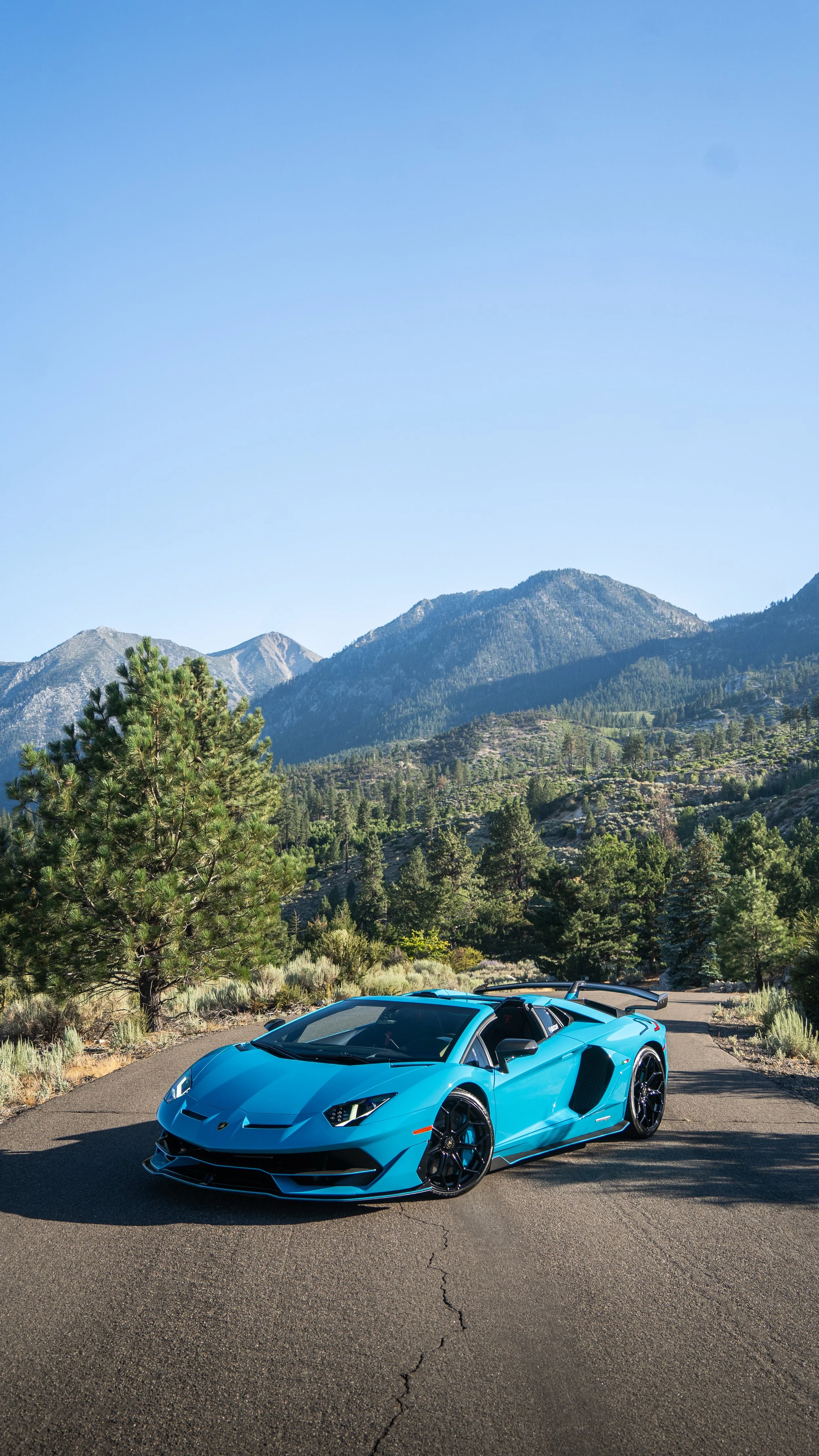 A bright blue sports car parked on a paved road in a mountainous area with green trees and clear blue sky.