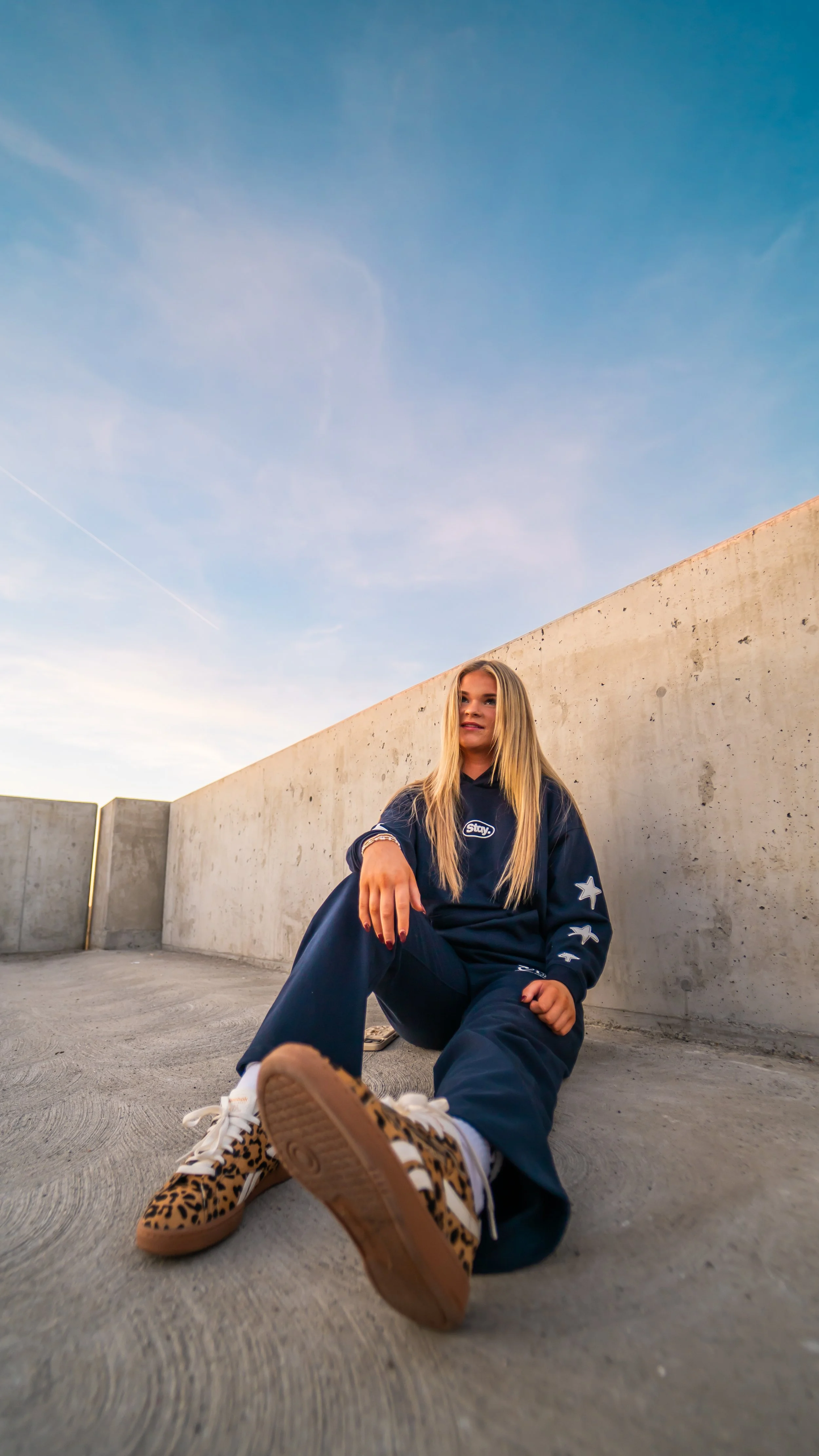 A young woman with long blonde hair sitting on the ground outdoors against a concrete wall at sunset, wearing a navy blue hoodie with patches, dark pants, and leopard print sneakers.