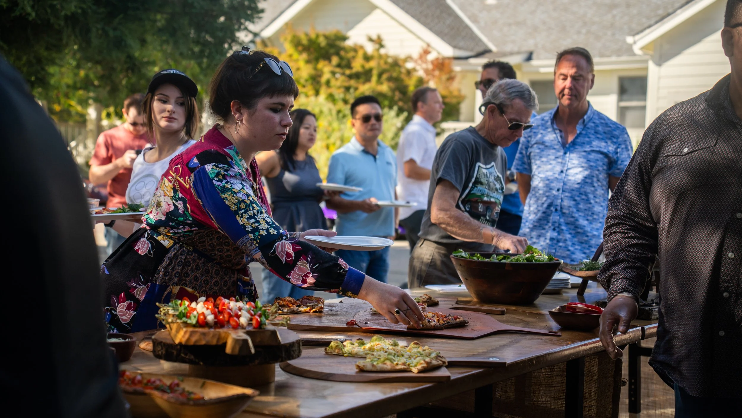 People serving and selecting food at an outdoor gathering or party with a variety of dishes on a long table.