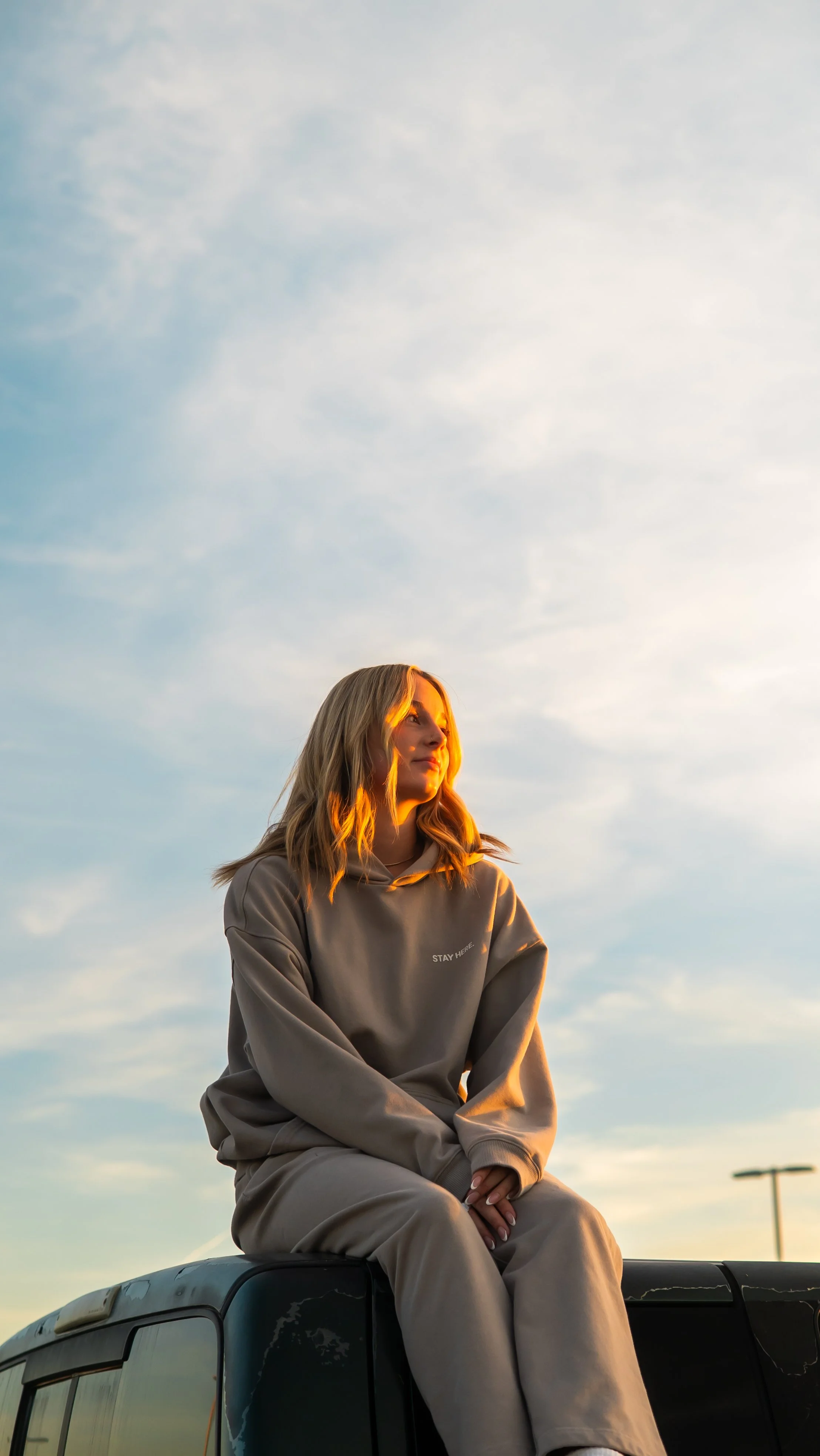 A young woman with blonde hair sits on top of a black vehicle, wearing a tan hoodie and matching pants, with a peaceful expression as she looks off into the distance during sunset, with a partly cloudy sky in the background.