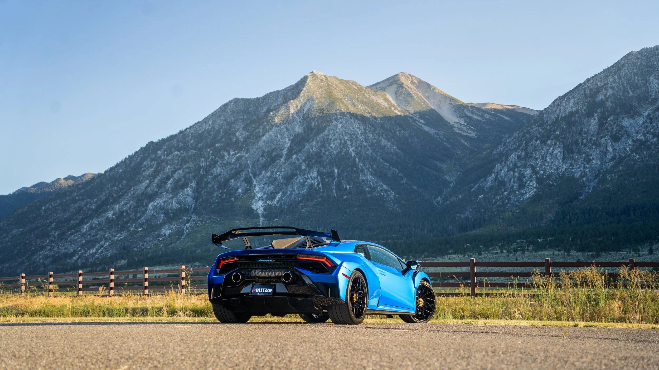 Blue Lamborghini sports car parked on a road with mountain peaks in the background and a wooden fence along the side of the road.