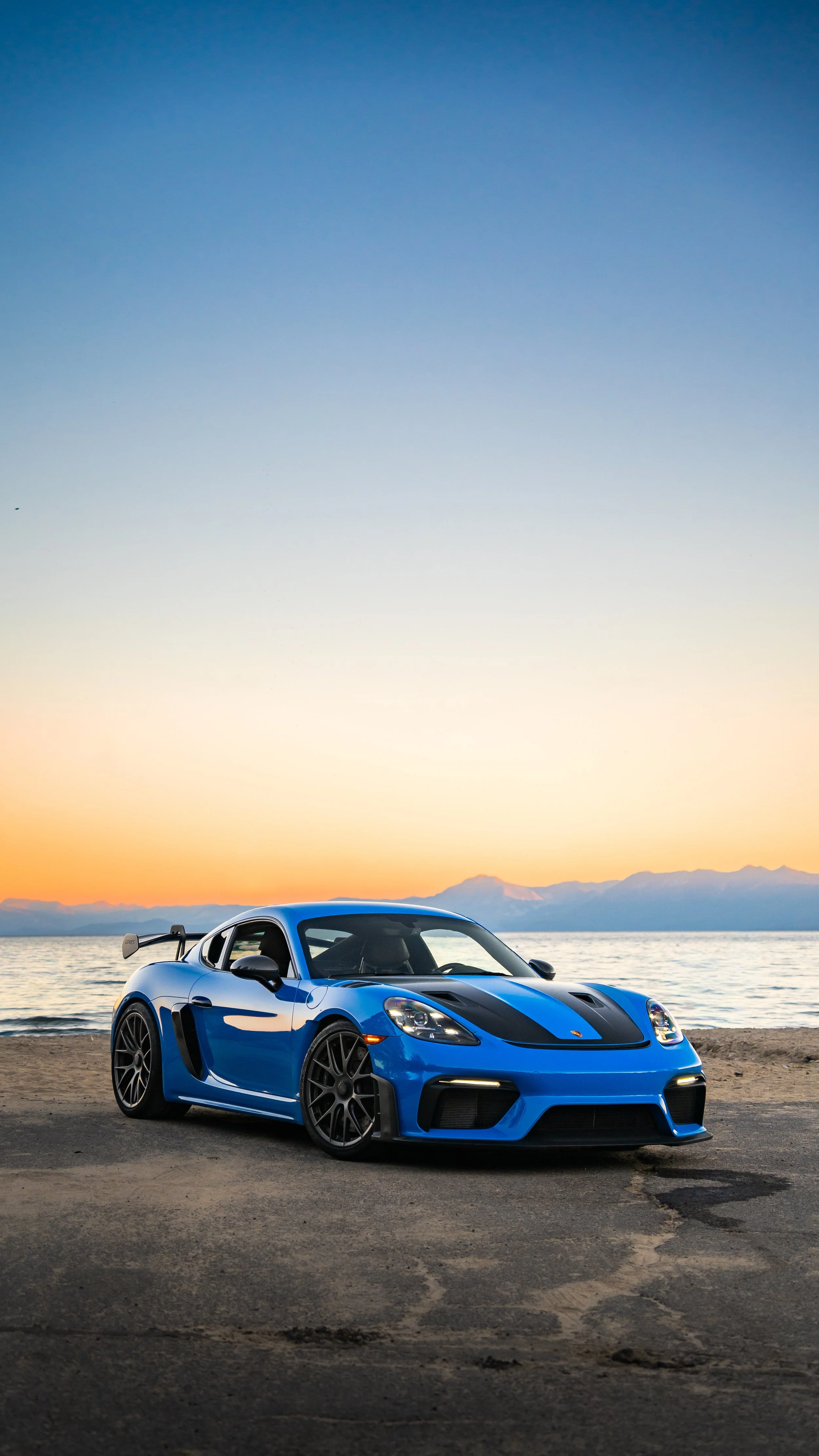 A blue sports car with black racing stripes parked on a beach at sunset, with mountains in the background.