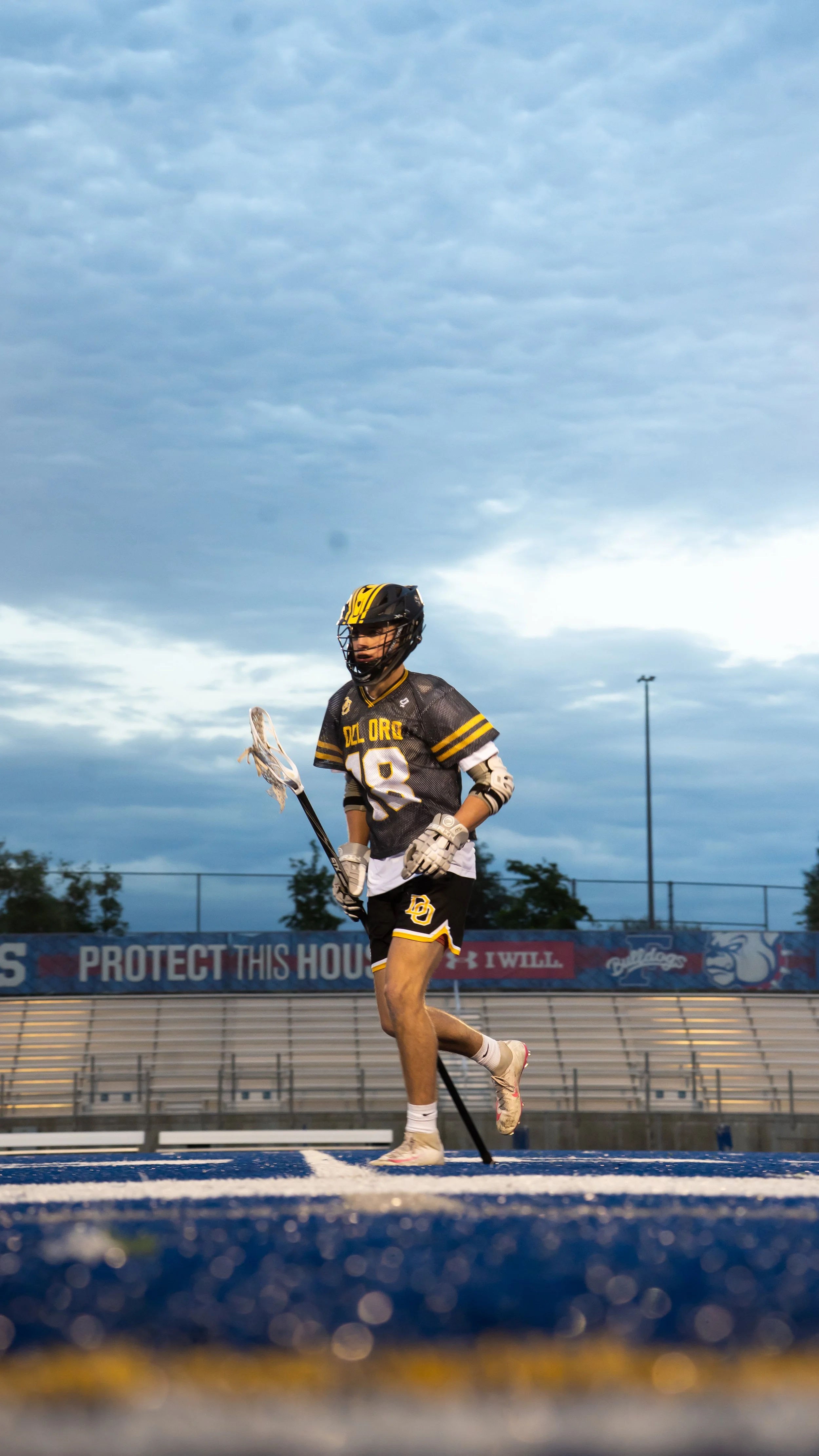 Lacrosse player in uniform with the number 18, running on the field with a lacrosse stick, sky with clouds overhead, empty bleachers in background.