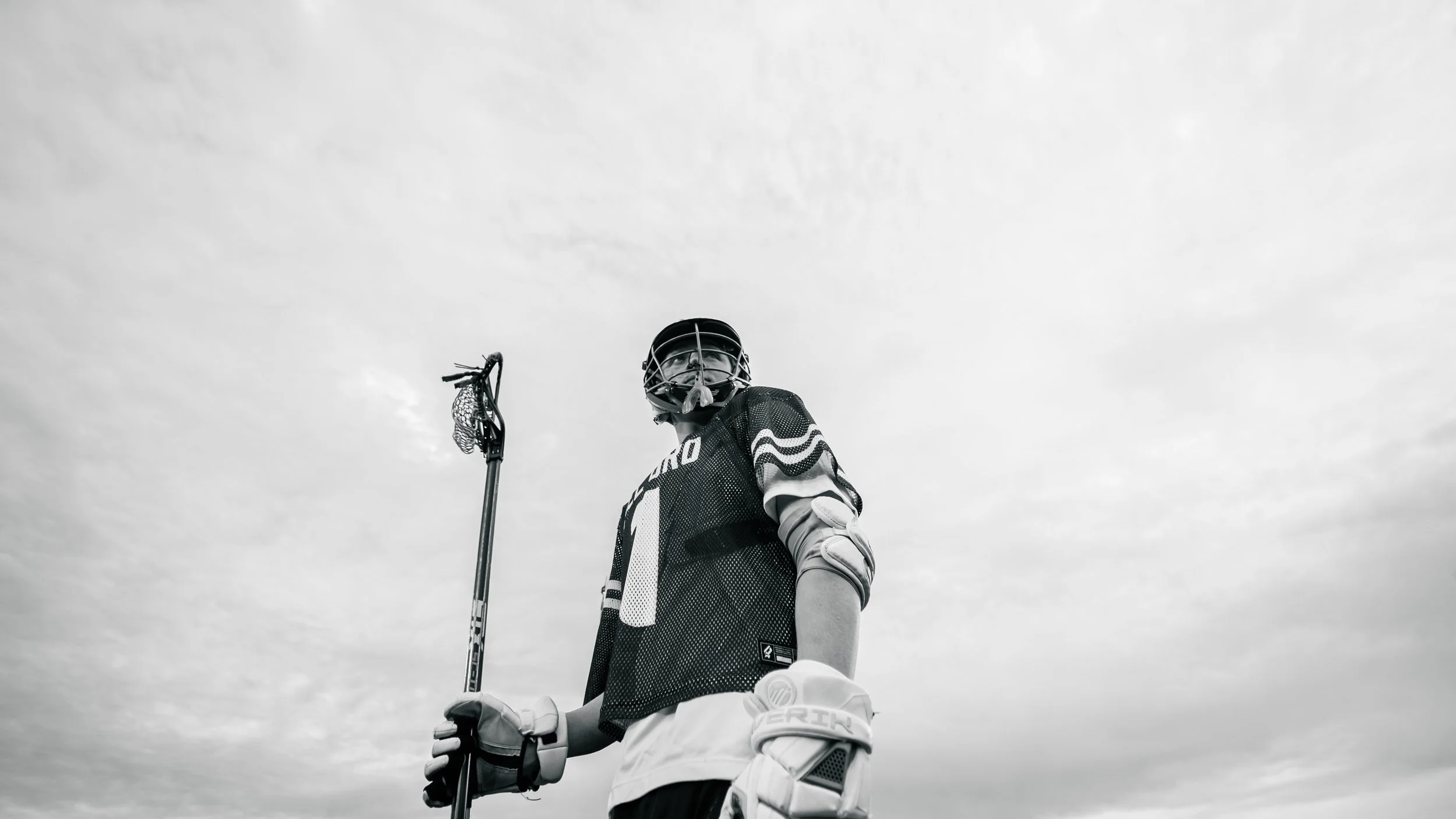 A black and white photo of a male field hockey player in uniform holding a field hockey stick, wearing a helmet, gloves, and sports gear, standing outdoors against a cloudy sky.