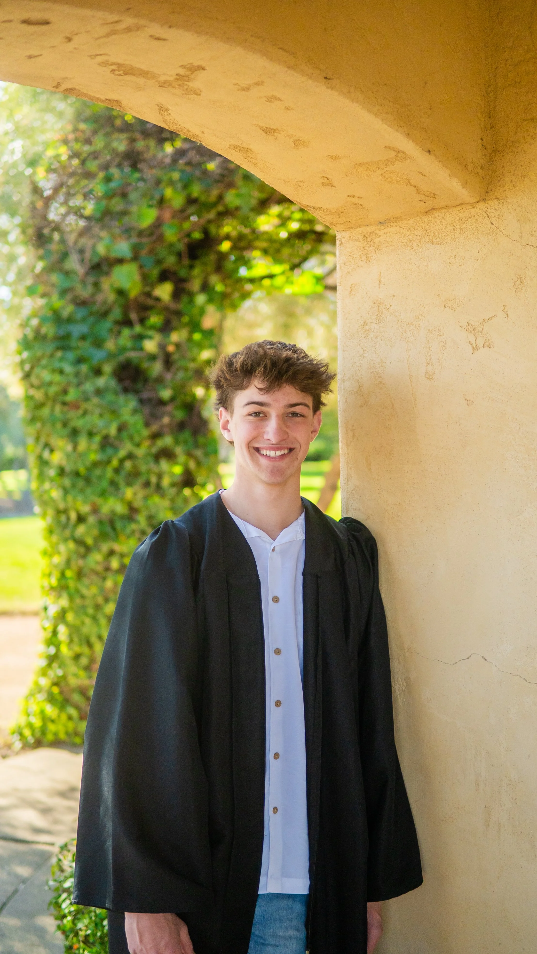 A young man in a graduation gown and white shirt smiling, standing beside a beige stone wall with greenery and trees in the background.