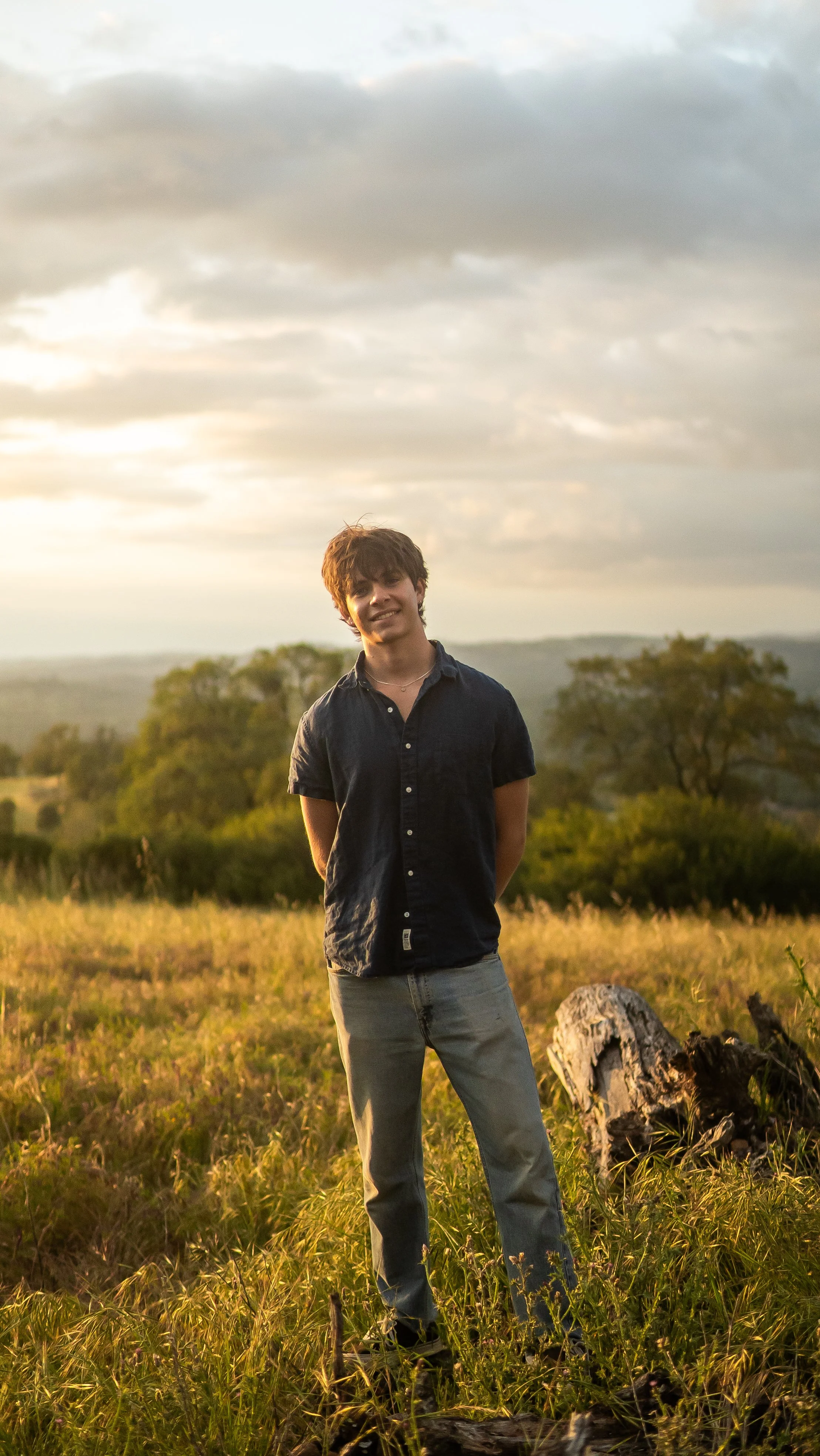 A young man standing in a grassy field during sunset, smiling and wearing a dark short-sleeve button-up shirt and light jeans with trees and distant hills in the background.