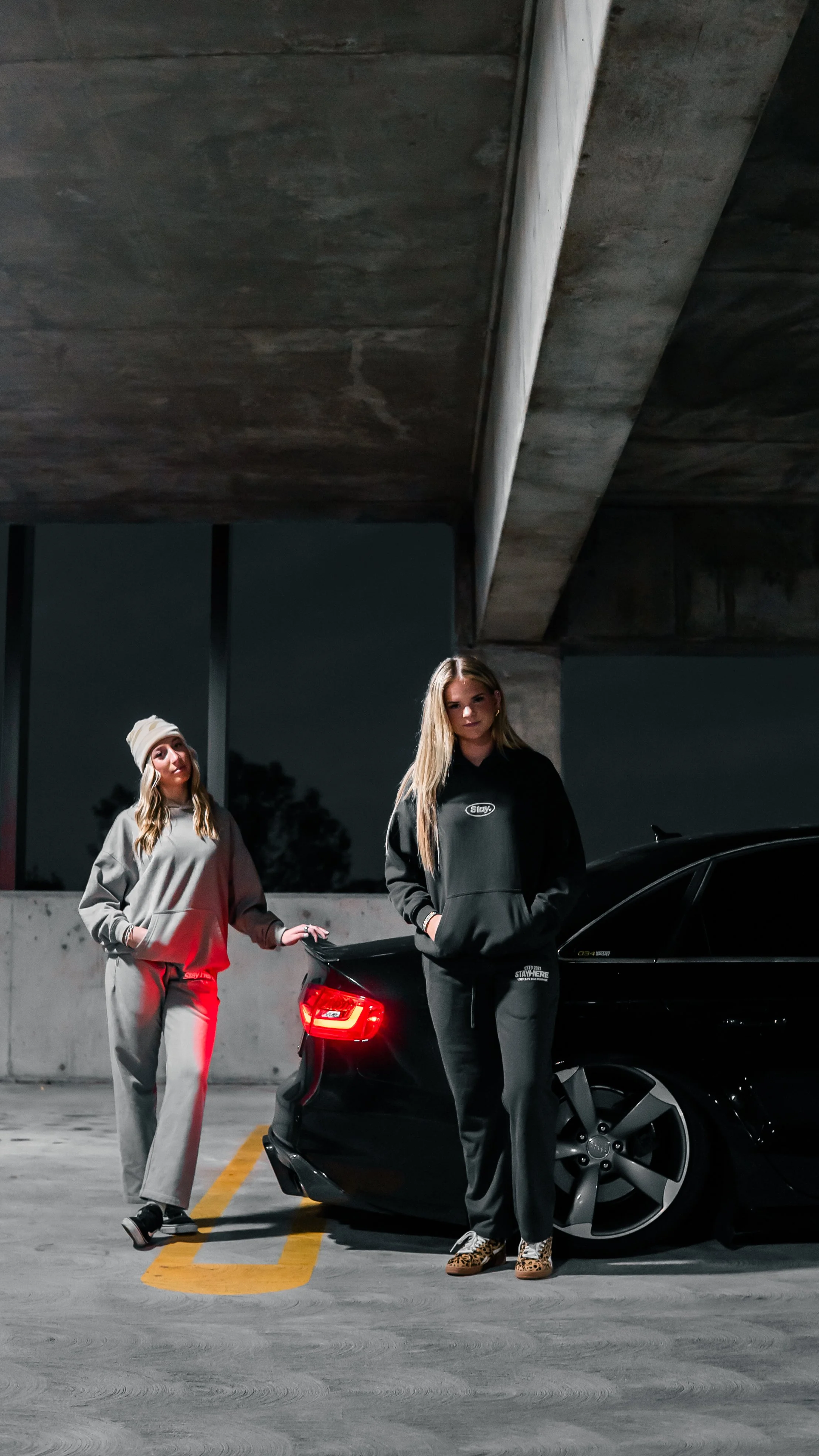 Two young women standing in a parking garage at night, next to a black sports car. One woman is wearing a beige hoodie, beige sweatpants, and a white beanie. The other woman is wearing a black hoodie and black pants with leopard print sneakers. Both 