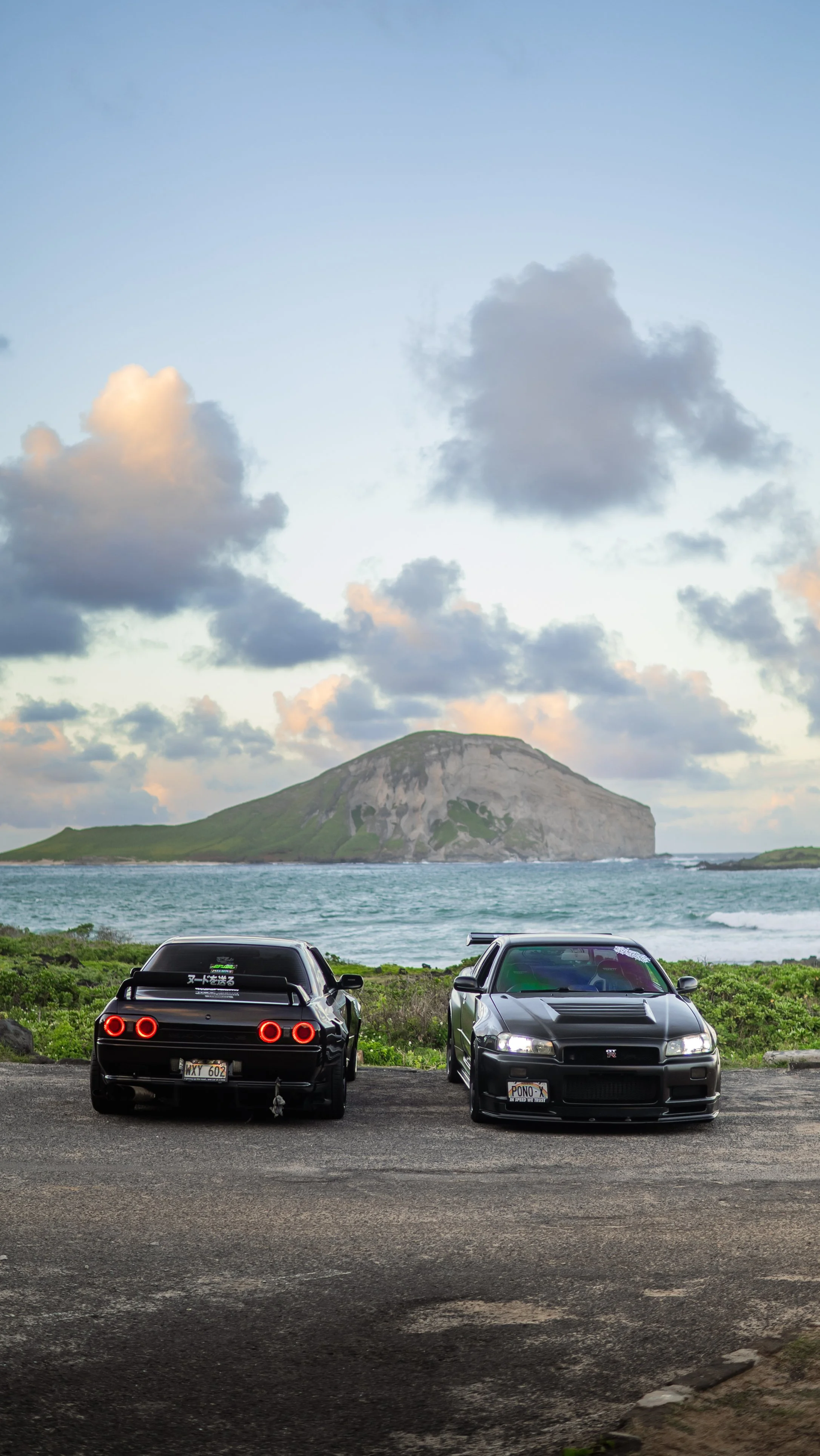 Two black sports cars parked on a gravel lot facing a body of water with a large island or mountain in the background under a partly cloudy sky.