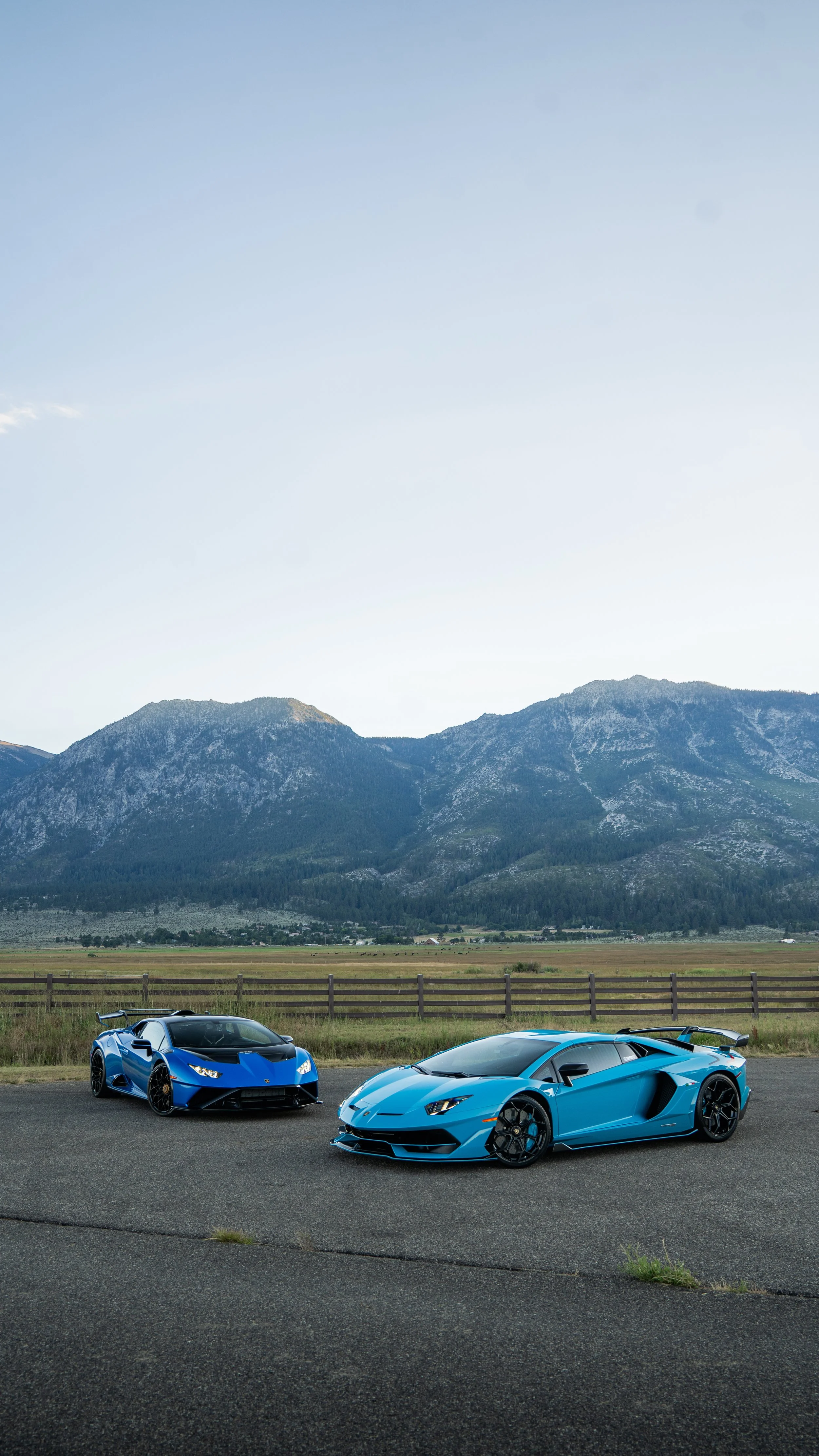 Two blue sports cars, a Lamborghini and a Ferrari, parked on the side of a road with mountains and a blue sky in the background.