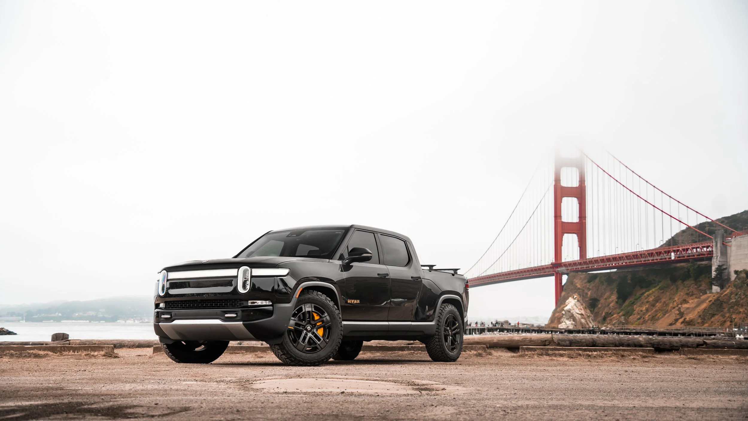 A black Rivian electric truck parked on a beach with the Golden Gate Bridge in the background.