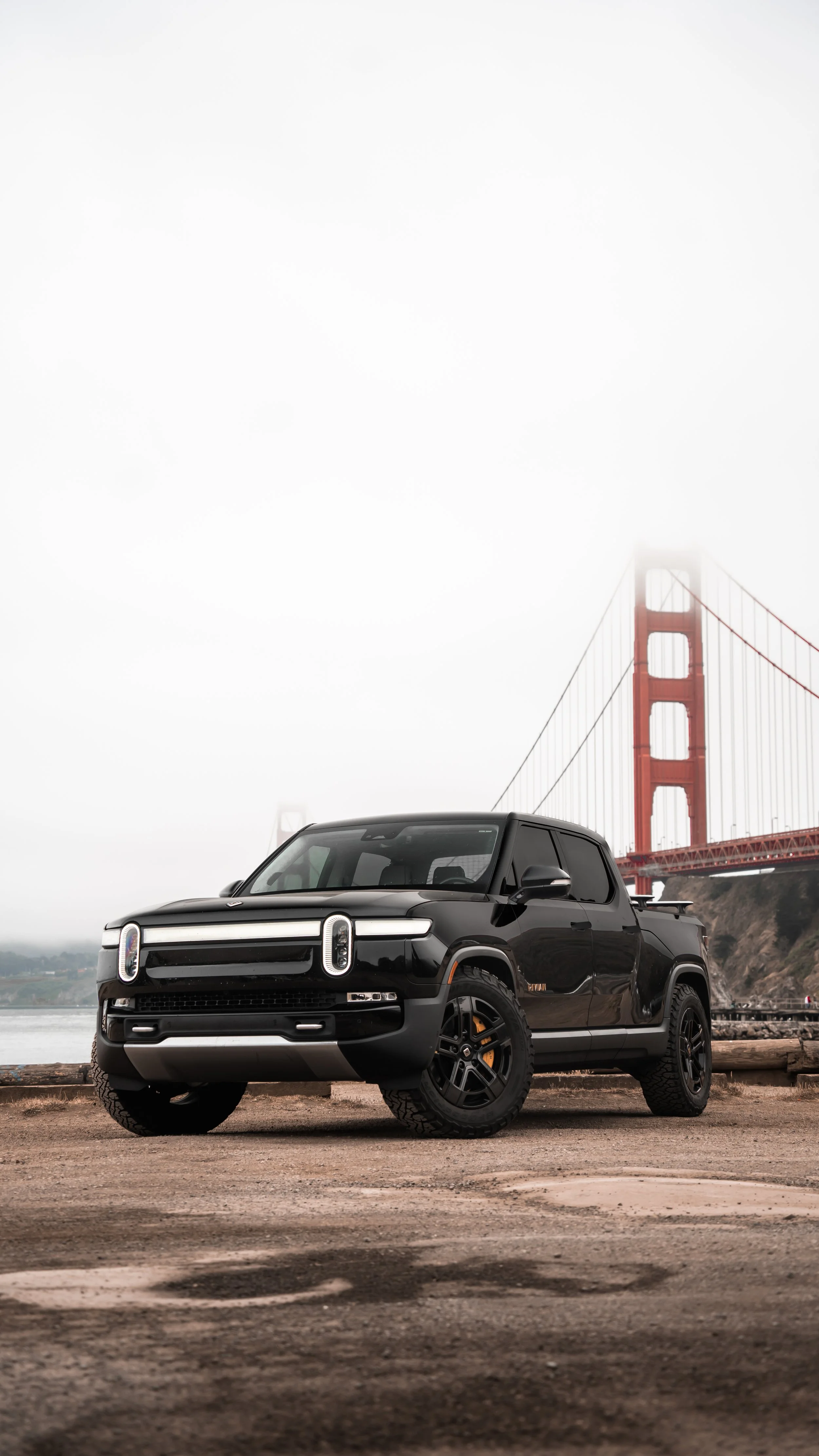 A black pickup truck parked on a dirt surface near the water with the Golden Gate Bridge in the background, partially obscured by fog.