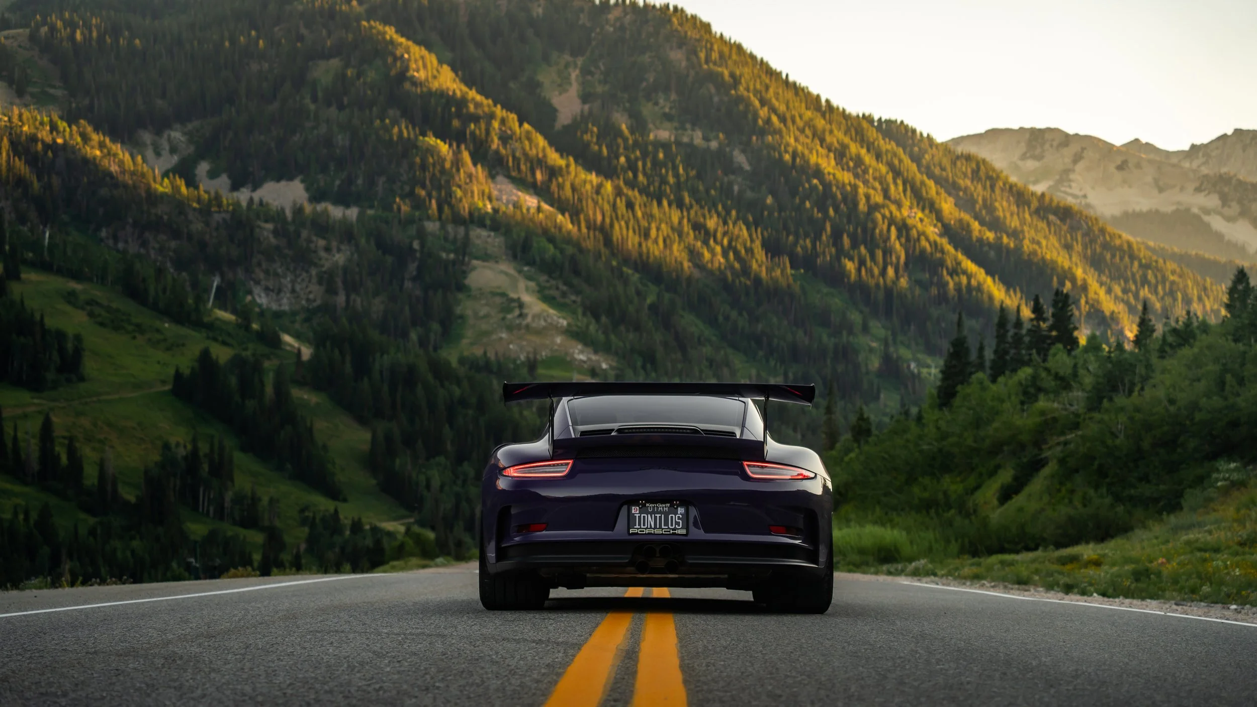 Back of a black sports car with a rear wing, driving on a two-lane mountain road surrounded by green forests and hills during sunset.