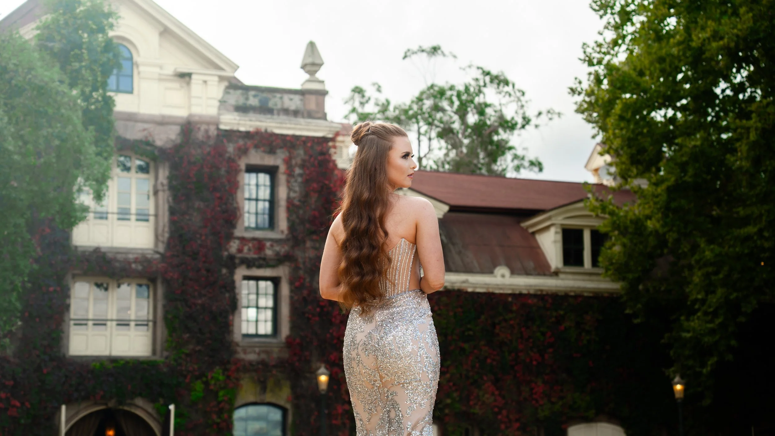 A woman with long wavy hair in a gown standing outdoors in front of a large house covered in ivy and trees.