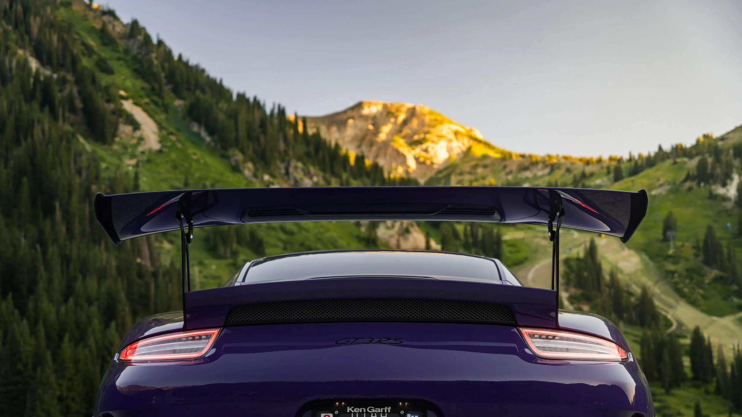Rear view of a purple Porsche GT3 RS sports car with a large rear spoiler, driving on a mountain road surrounded by green trees and hills at sunset.