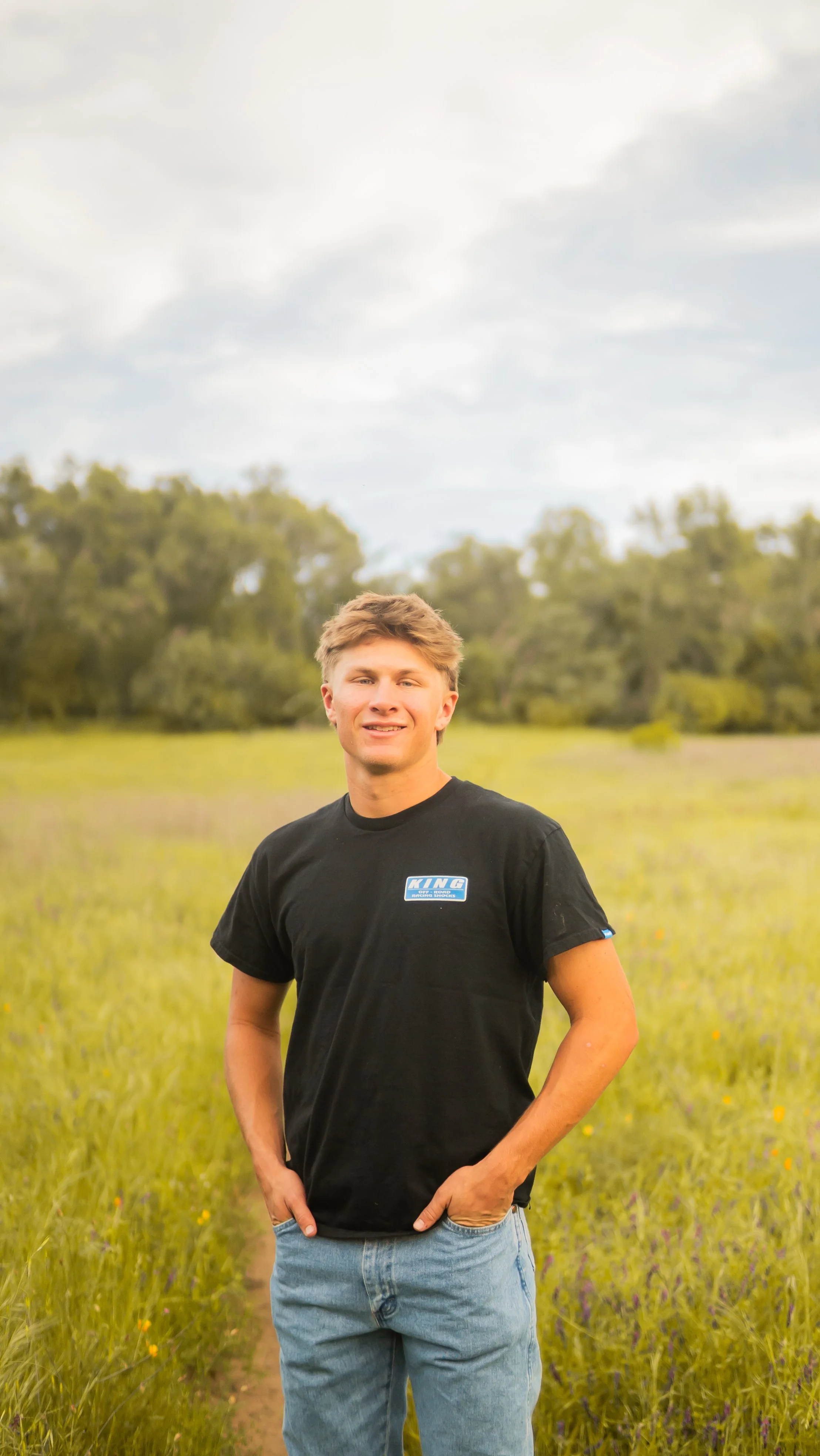 A young man standing in a grassy field with trees in the background, wearing a black t-shirt with a blue and white logo that reads 'KING' and blue jeans.