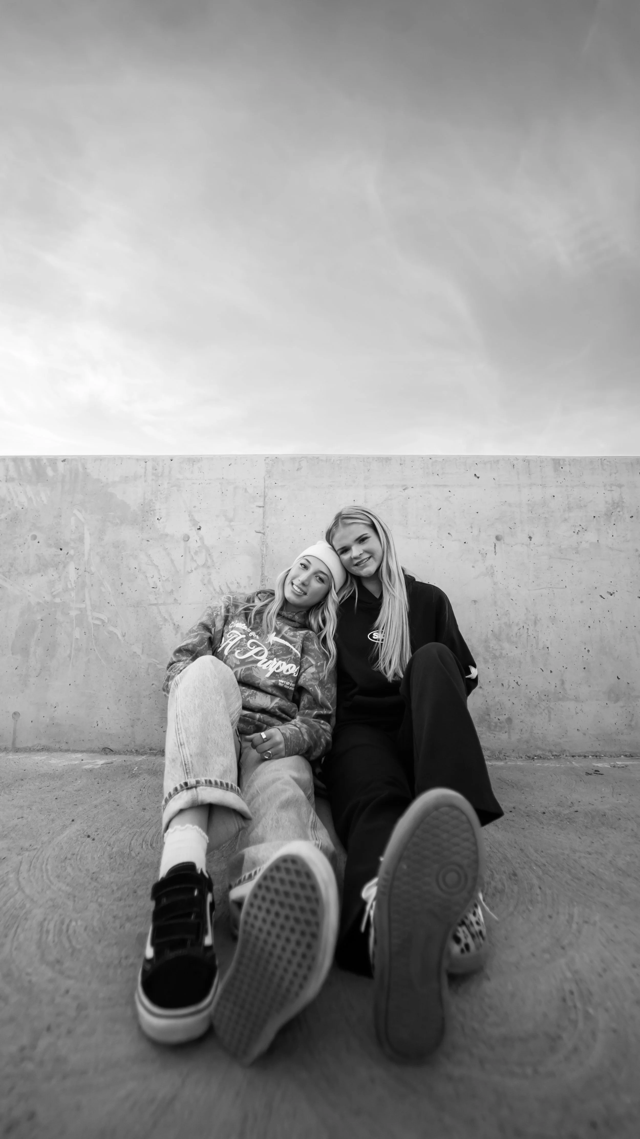Two young women sitting on the ground against a concrete wall, smiling and looking at the camera. One is wearing a hoodie and the other is wearing a sweatshirt.
