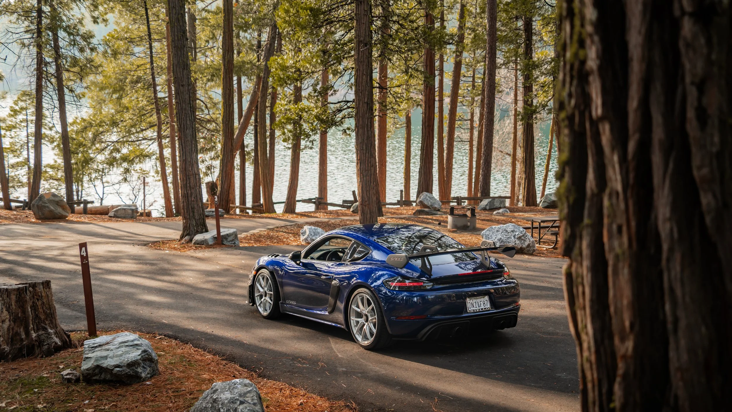 A black sports car parked on a paved area near a lakeshore, with tall trees and large rocks surrounding the scene.