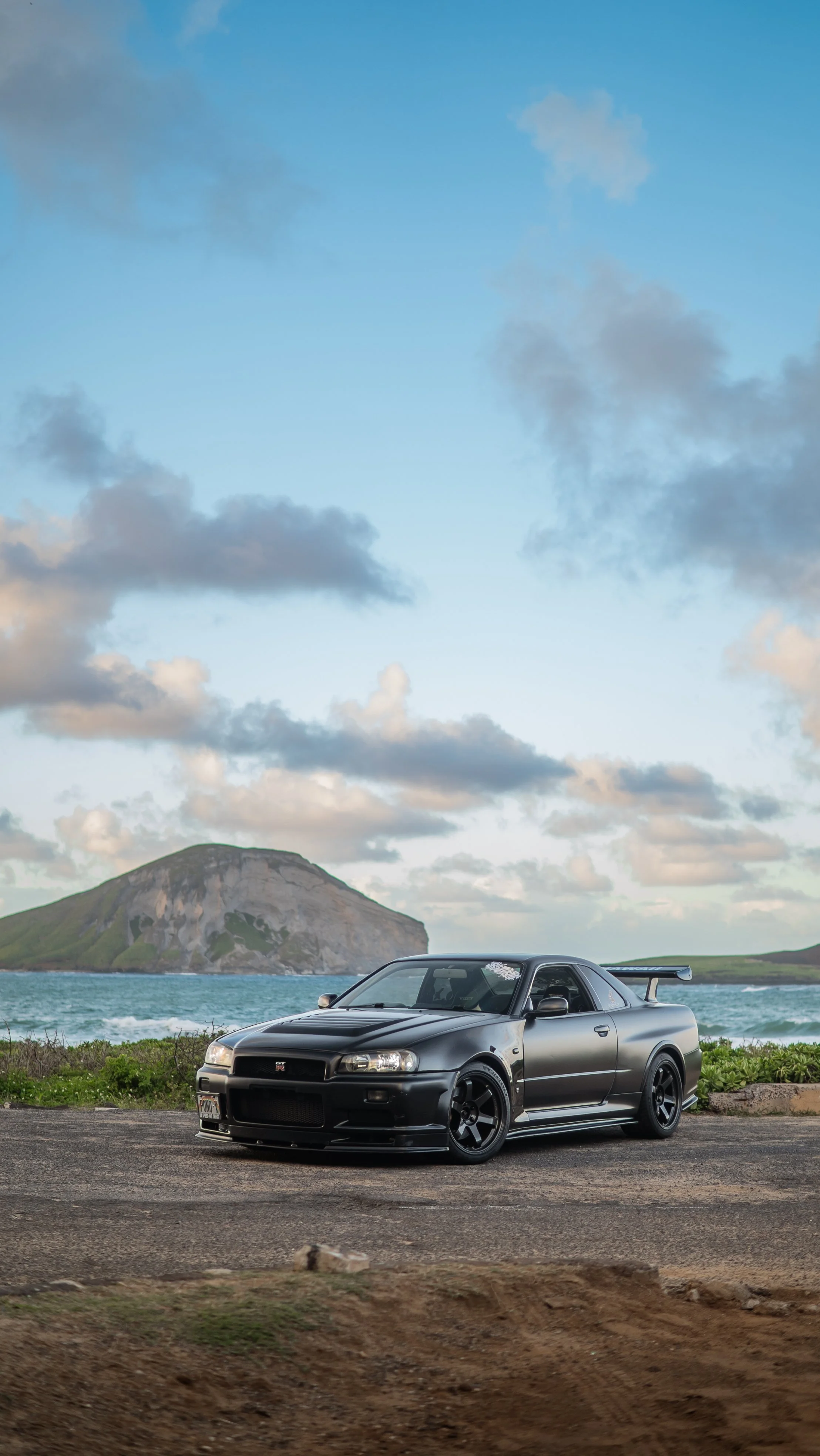 Black sports car parked near the ocean with a small island and mountain in the background, under partly cloudy sky.