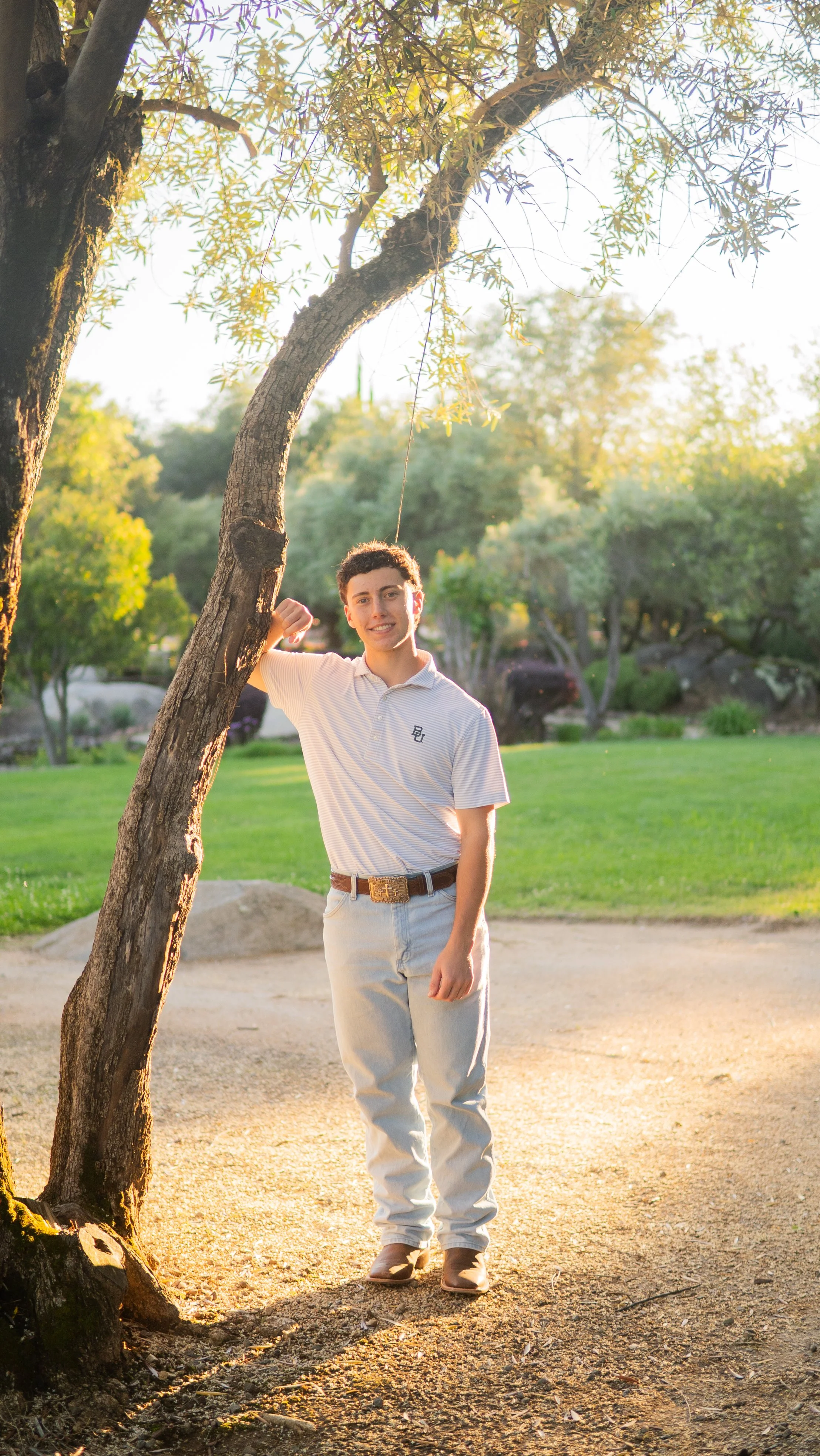 A young man with curly hair and fair skin standing outdoors near a tree with a swing, during golden hour sunset, surrounded by greenery and trees, wearing a light striped polo shirt, beige pants, and cowboy boots.