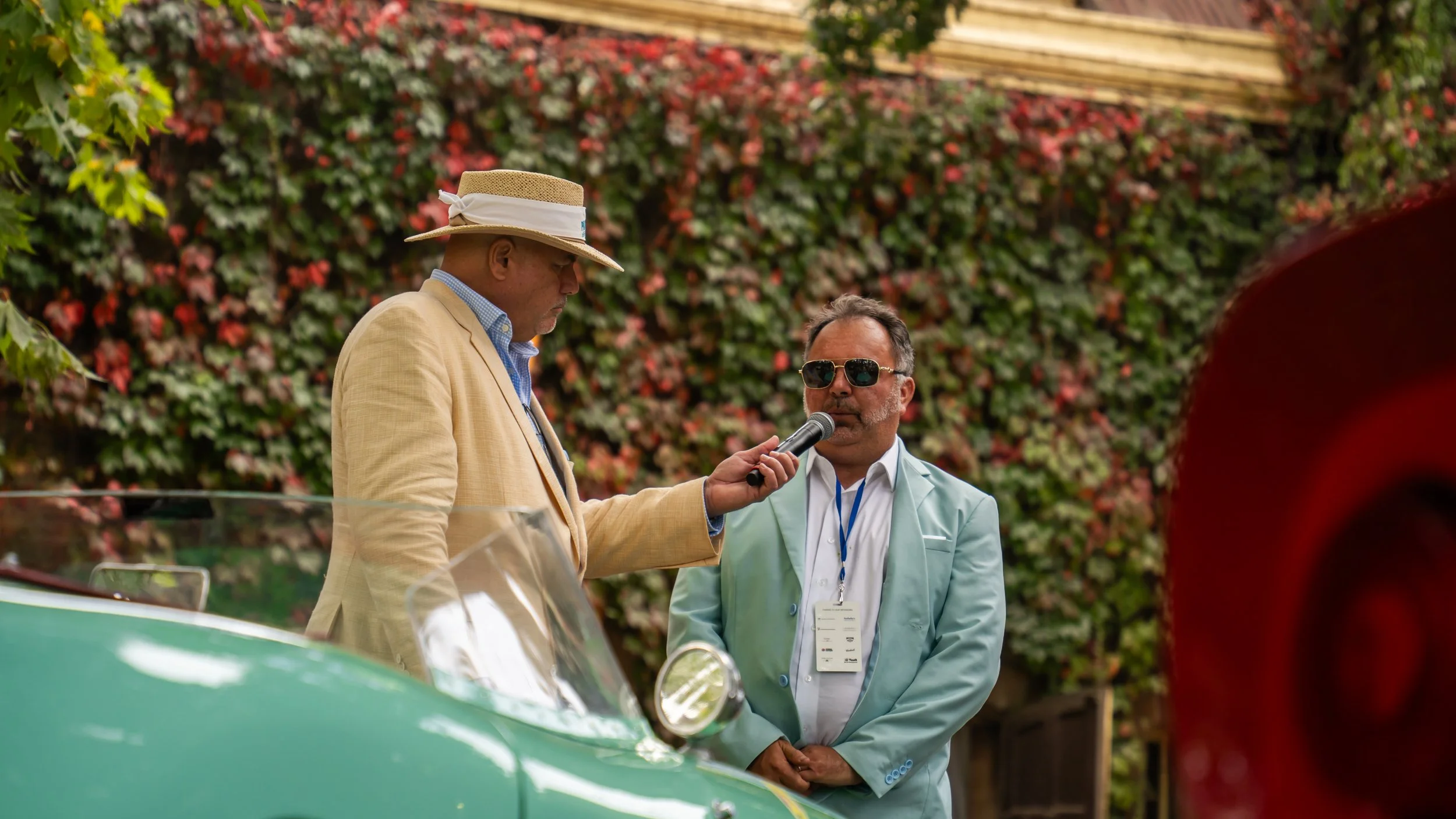 Man in a beige jacket and straw hat being interviewed by another man in a mint green jacket and sunglasses, with a background of pink and green plants.