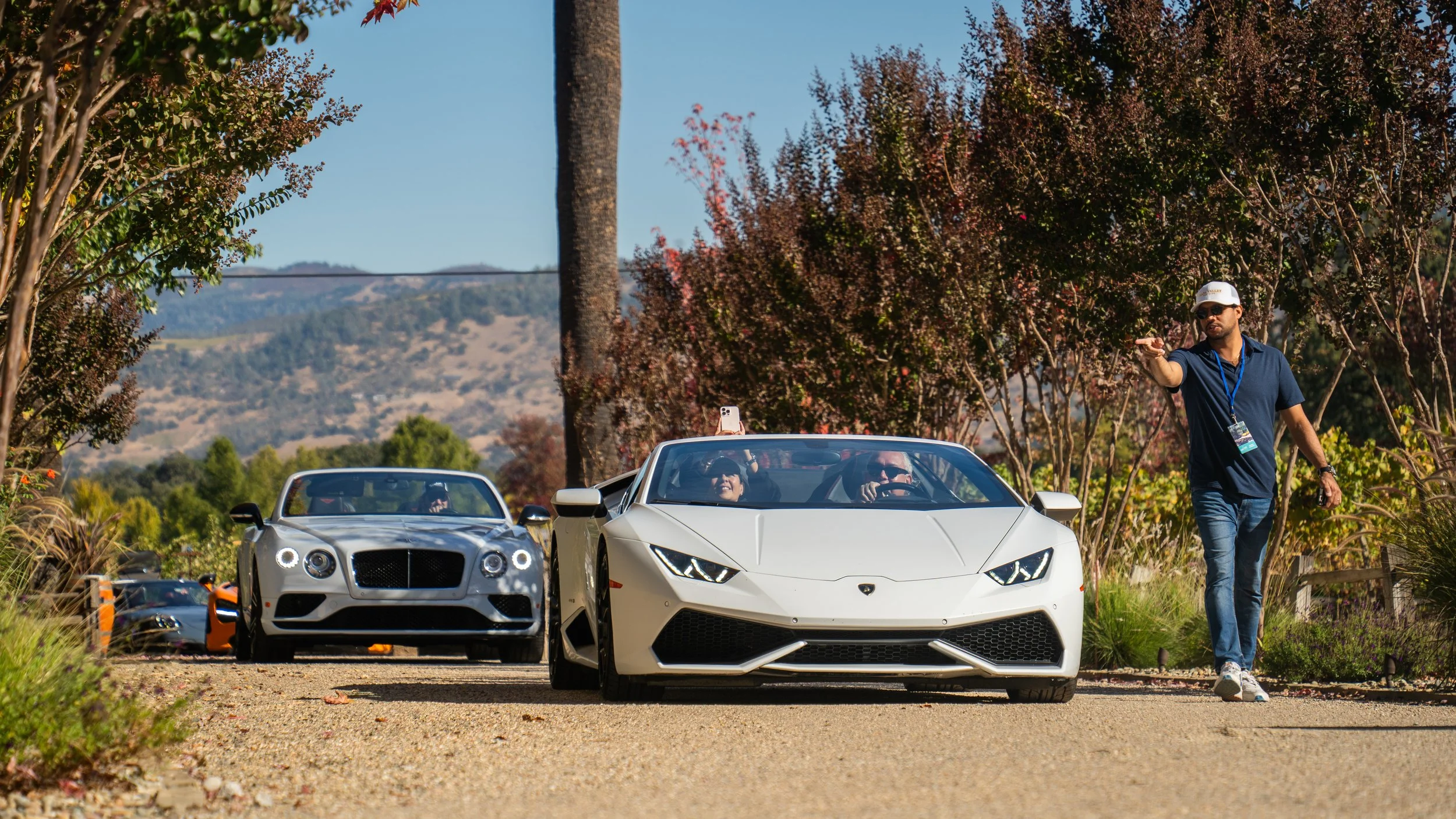 A man walking in front of a white Lamborghini sports car, with a woman and a man inside the car, and another white sports car behind them. The setting appears to be a sunny outdoor area with trees and mountains in the background.