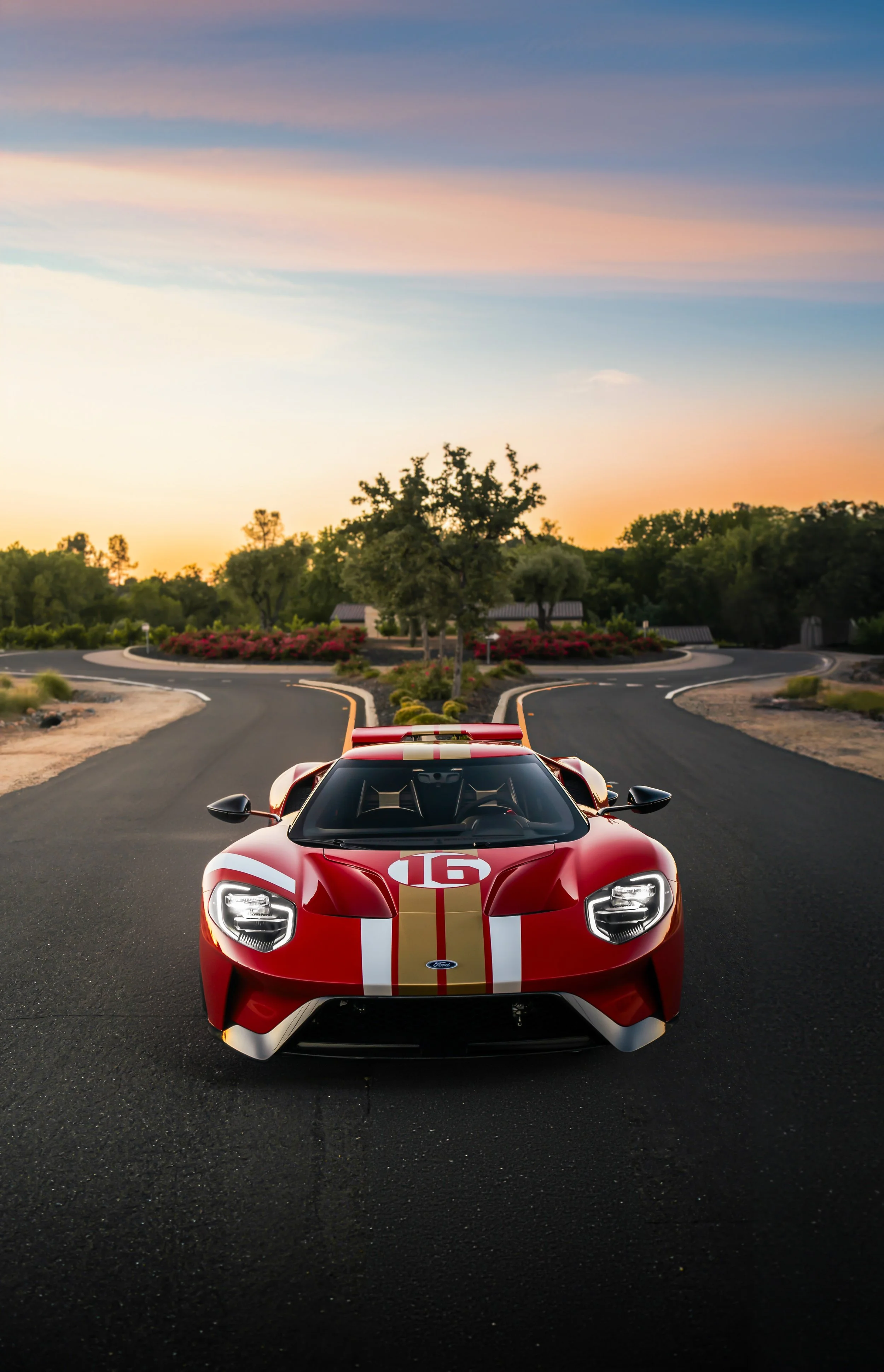 Red sports car with racing stripes on a paved driveway at sunset, with landscaped bushes and trees in the background.