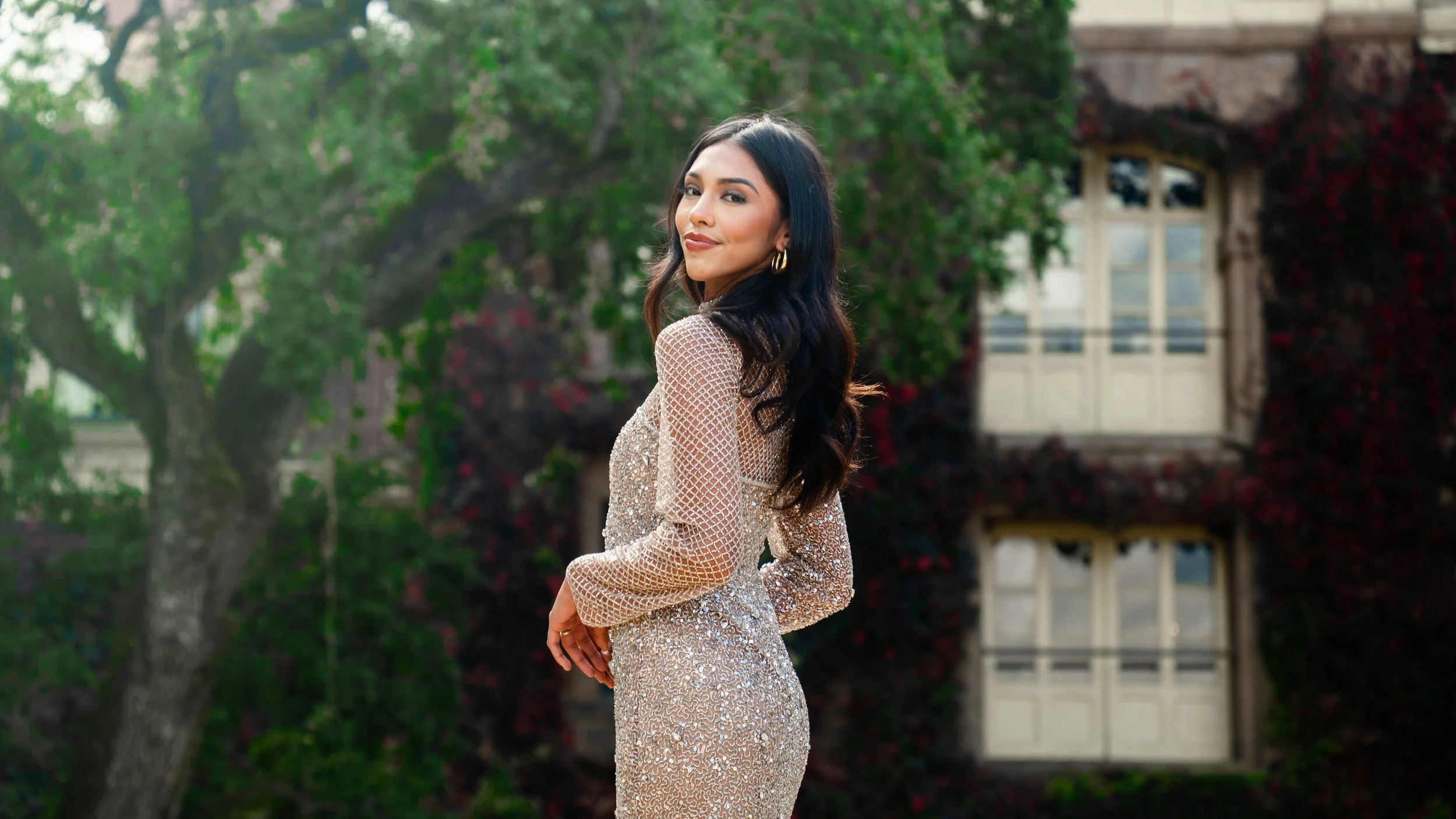 A woman with long dark hair in a gold dress standing outdoors with greenery and a building in the background.