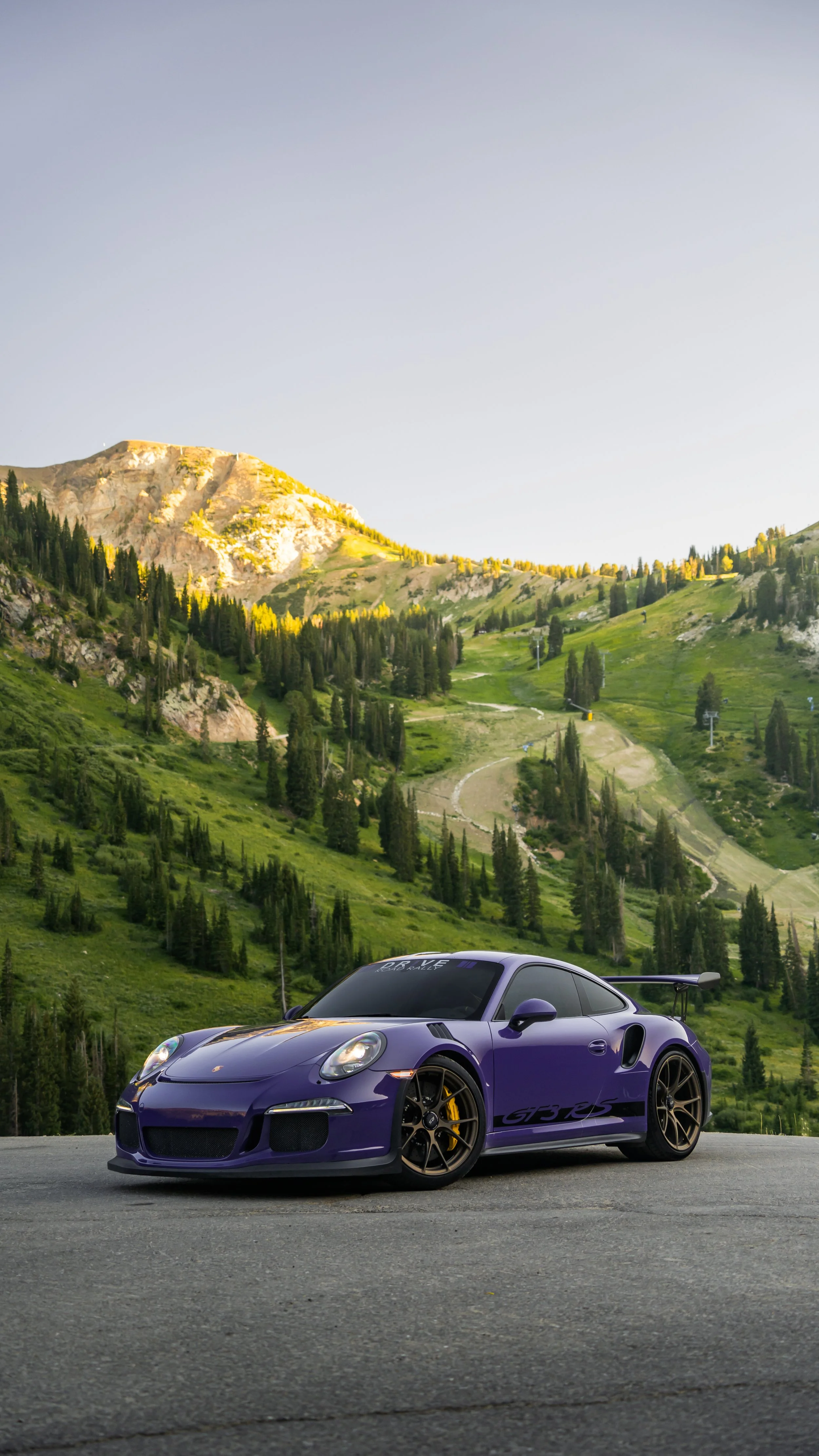 A purple Porsche 911 GTS RS parked on a mountain road with green hills, trees, and a clear sky in the background.
