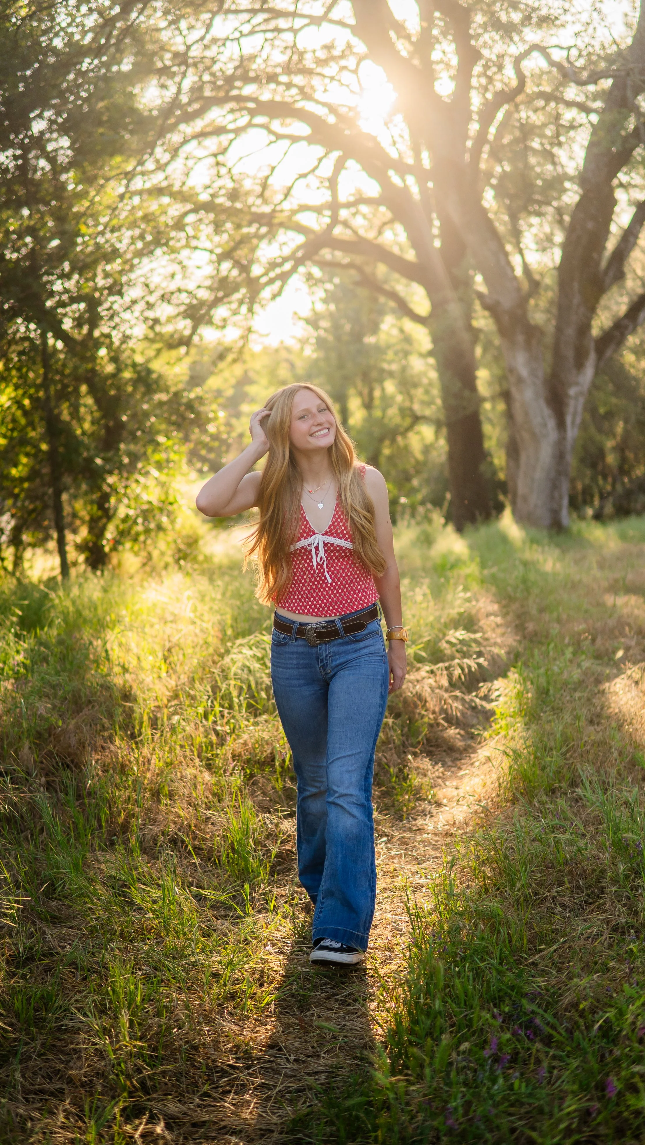 A young woman with long red hair smiling and walking down a dirt trail in a wooded area during sunset. She is wearing a red sleeveless top with white patterns, blue jeans, and sneakers, with sunlight filtering through the trees.